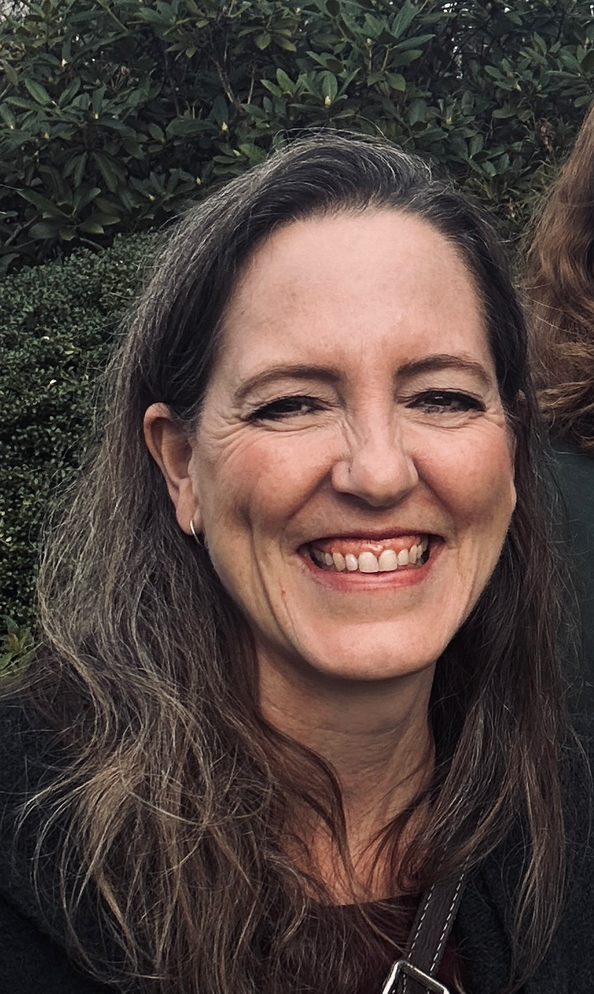 A client of Andrea Tessier, IFS coach. Close-up of a smiling woman with long, wavy brown hair, wearing a small hoop earring, and standing outdoors with green bushes in the background.