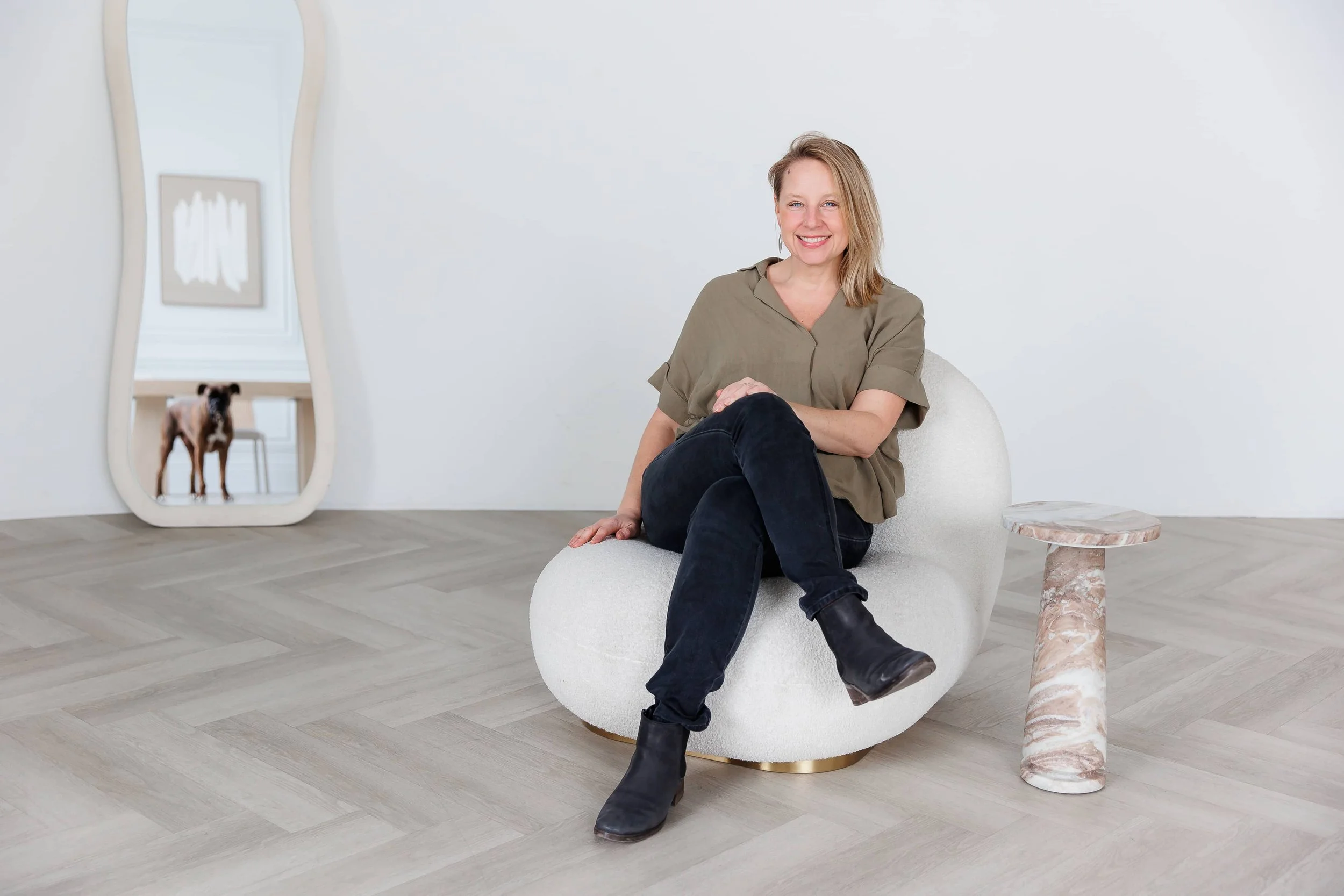 Andrea Tessier, leadership women's coach, with blonde hair sitting on a white round chair, smiling, wearing a tan shirt and black pants, in a minimalist room and a dog visible through the mirror.