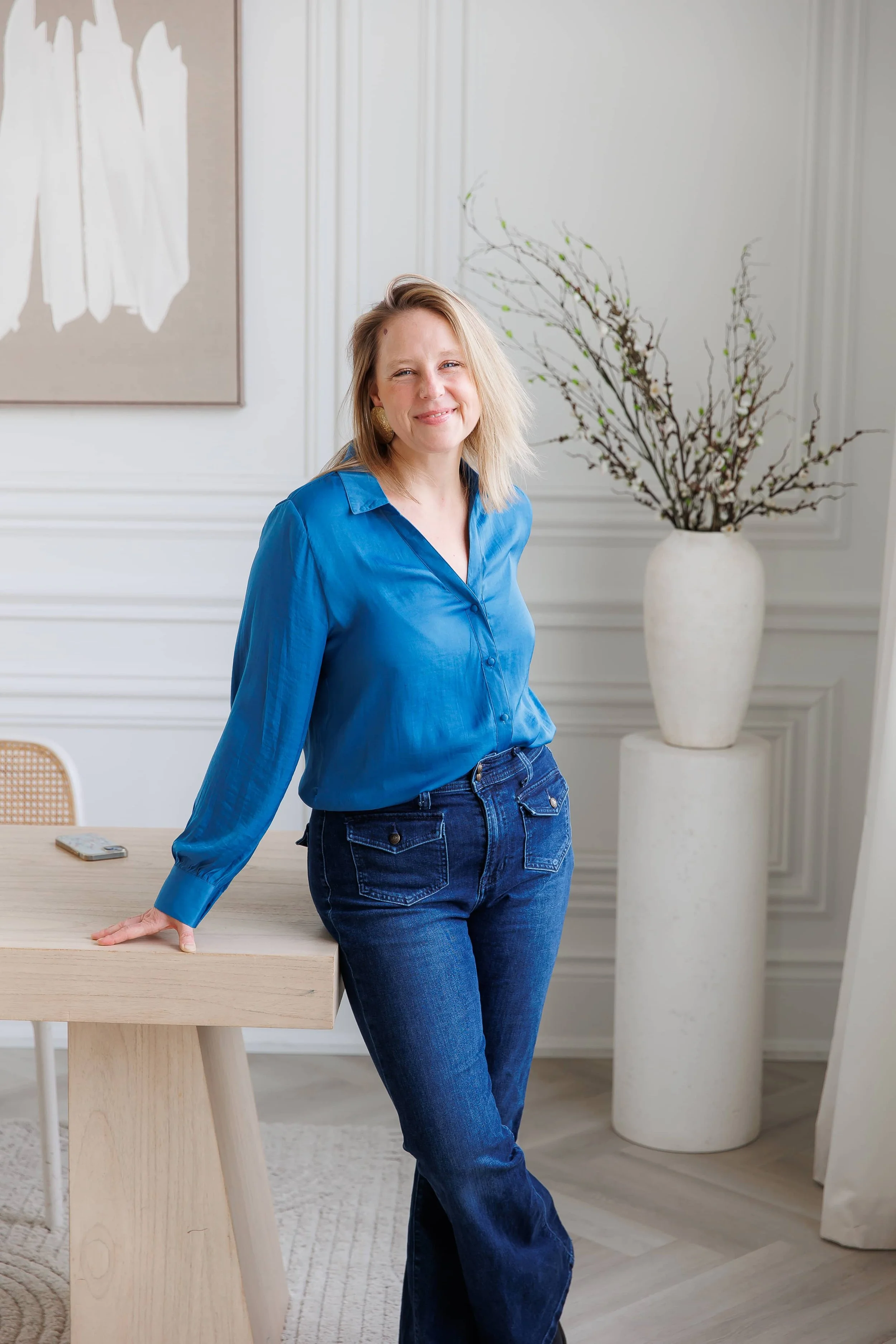 Andrea Tessier, a women's life coach, with shoulder-length blonde hair wearing a blue blouse and dark blue jeans, standing in a bright, modern room with white walls. She is leaning against a wooden table, smiling, and looking at the camera.