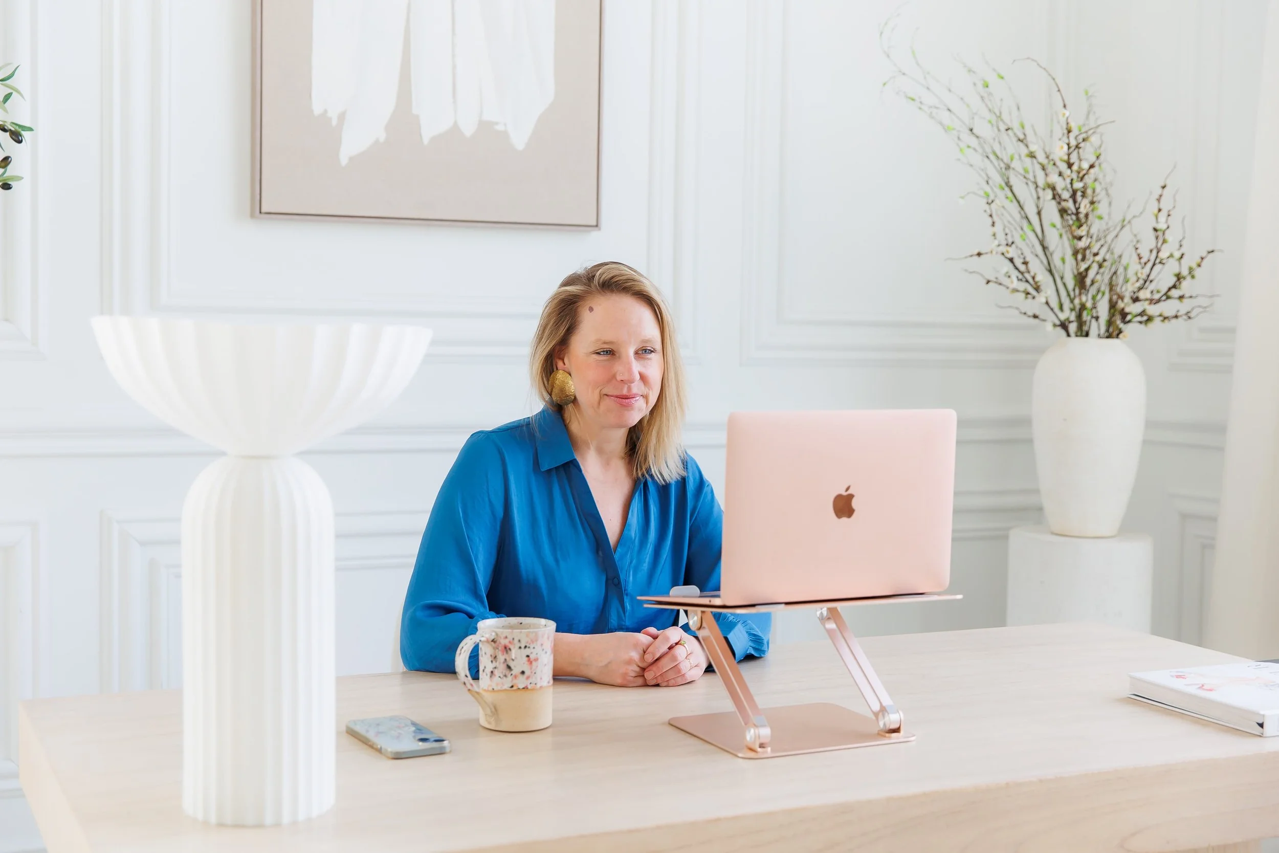 Andrea Tessier, coach for high achieving women, with blonde hair, wearing a blue shirt, sitting at a light-colored wooden desk in a bright, white room, working on a pink laptop. There are decorative vases and a floral mug on the desk.