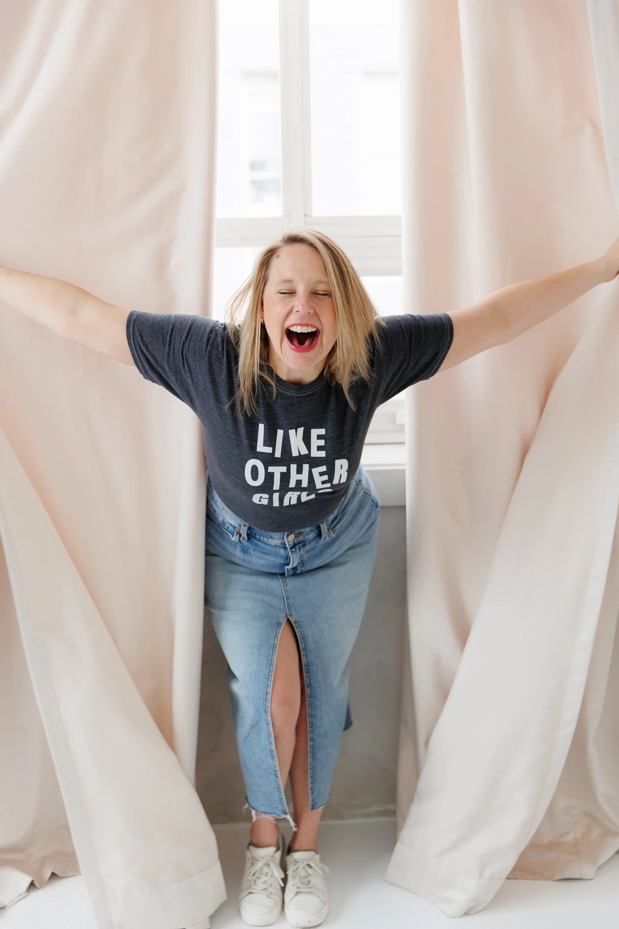 A woman with blonde hair standing between beige curtains, stretching her arms out wide and leaning forward with her eyes closed and mouth open in a joyful expression. She's wearing a dark grey T-shirt that says 'LIKE OTHER GIRLS' and a ripped denim skirt, with white sneakers.