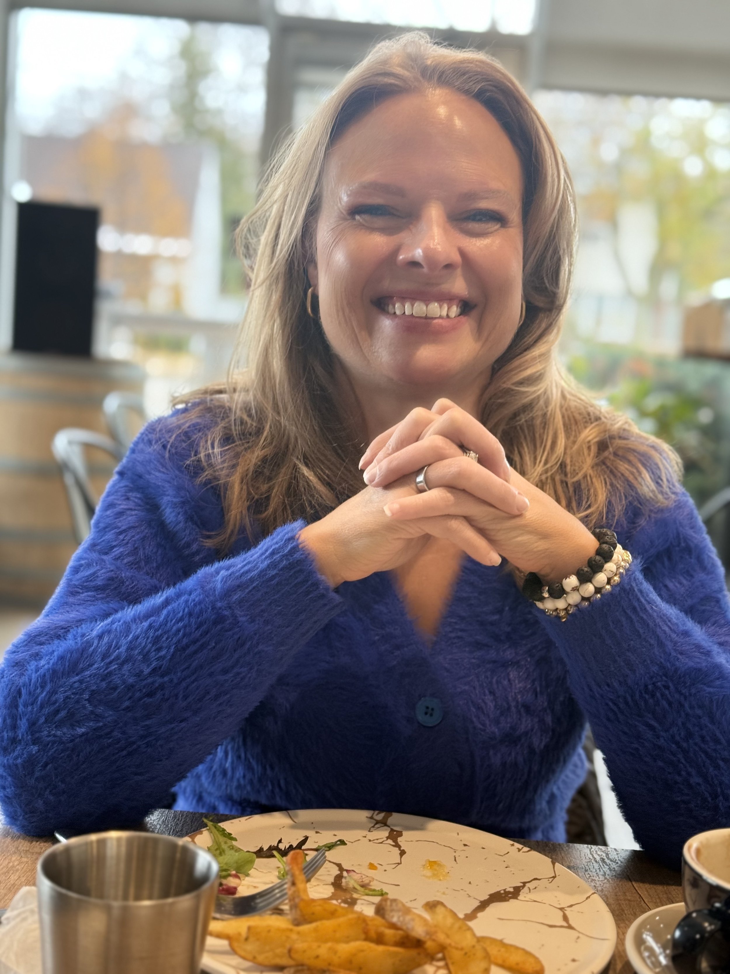 A client of Andrea Tessier, toronto-based coach. A woman with long hair wearing a blue fuzzy sweater, smiling with hands clasped, sitting at a table with food and drinks in a bright indoor setting with large windows and blurred background.