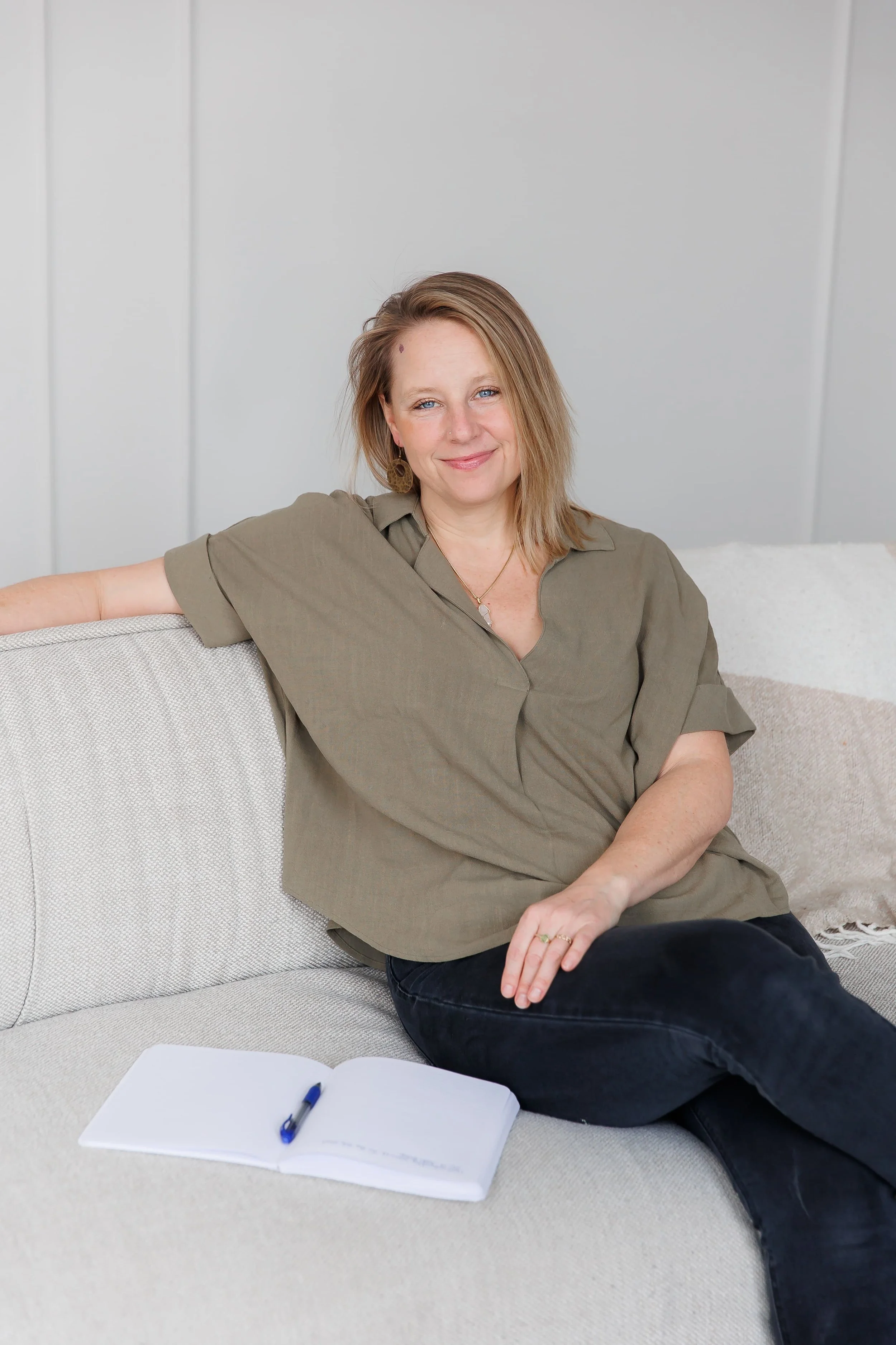 Andrea Tessier, a women's coach with shoulder-length hair, sitting on a light-colored couch in a room with light gray walls. She is smiling at the camera with a relaxed posture. An open notebook and pen are on the couch in front of her.