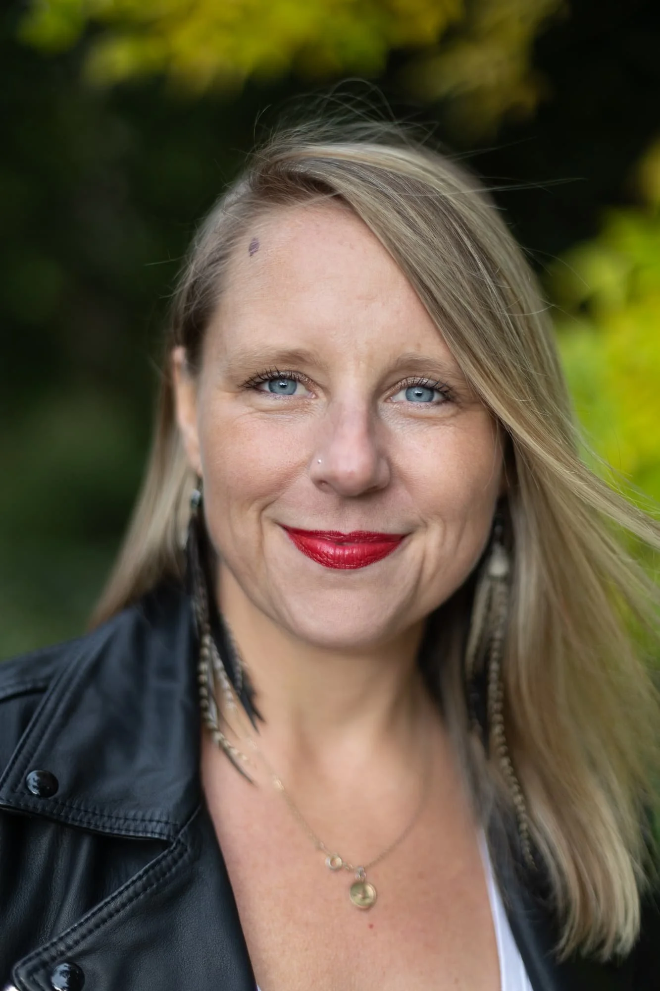Andrea Tessier, IFS coach, with blonde hair, blue eyes, and red lipstick smiling outdoors, wearing a black leather jacket and layered necklaces.