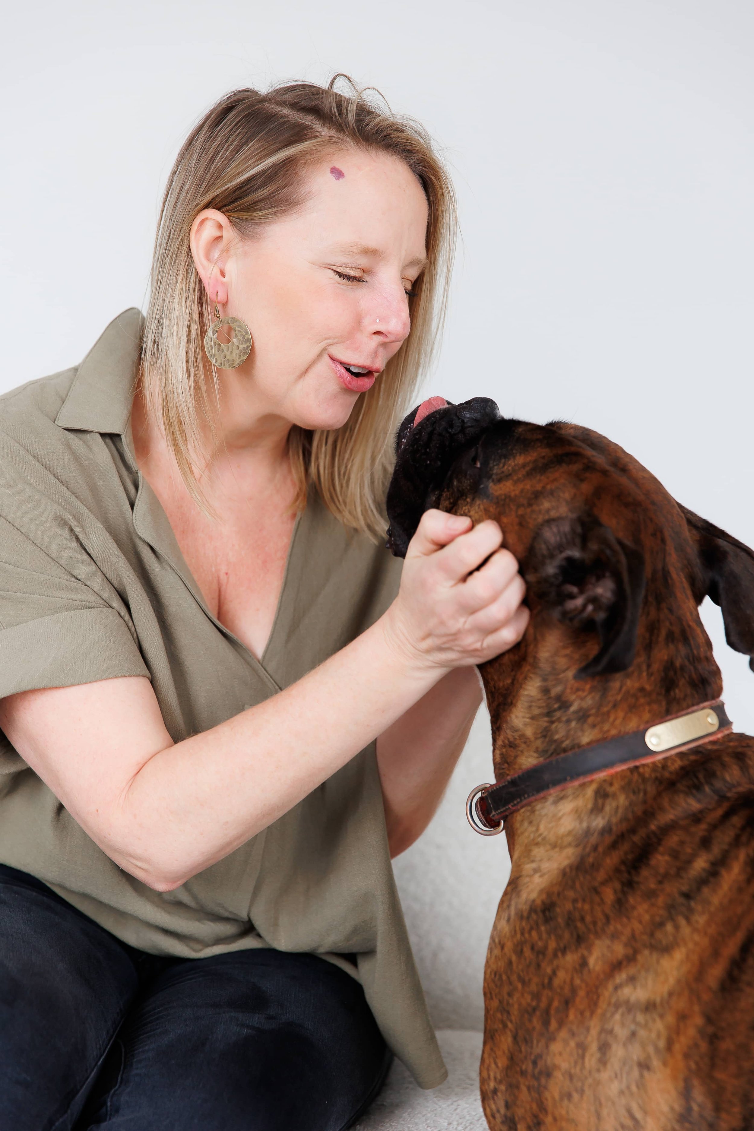 Andrea Tessier, an Internal Family Systems coach, with blonde hair and earrings interacts with a large brown and black brindle dog, both facing each other with the woman smiling.