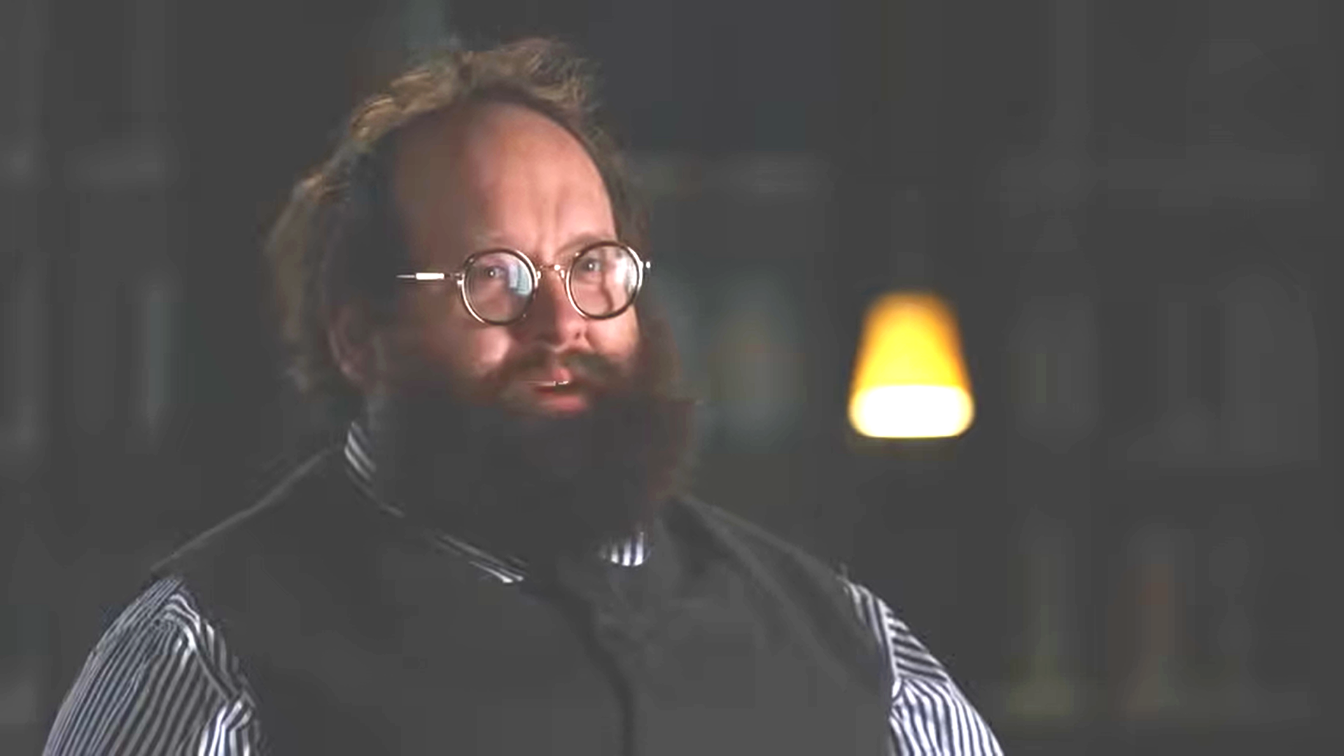 Dell J. Rose with glasses, curly hair, and a beard wearing a striped shirt and black vest, sitting in a dimly lit room with a yellow lamp in the background.