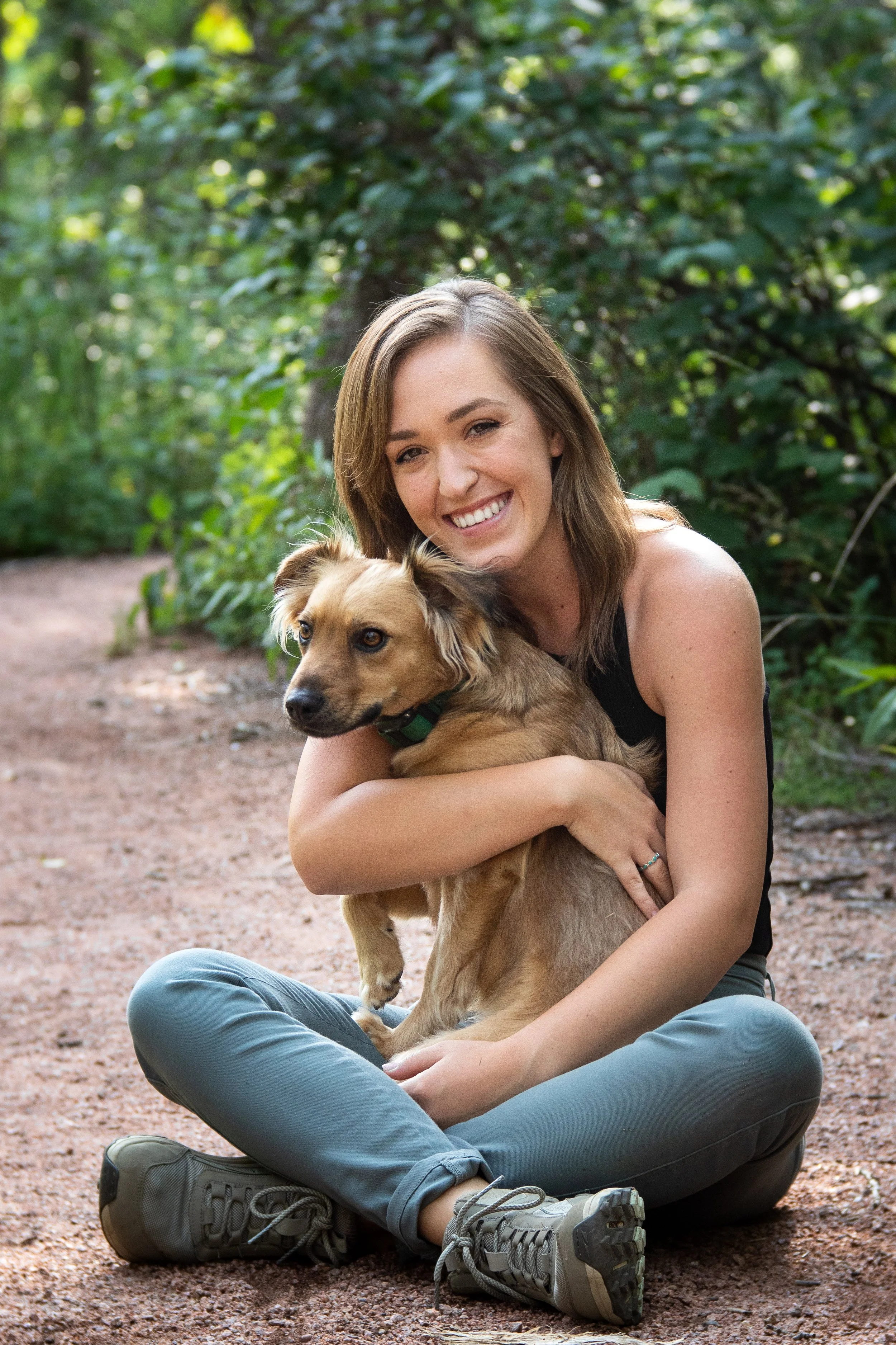 A young woman sitting cross-legged on a dirt trail hugging a small brown dog with a green collar, surrounded by lush green trees.