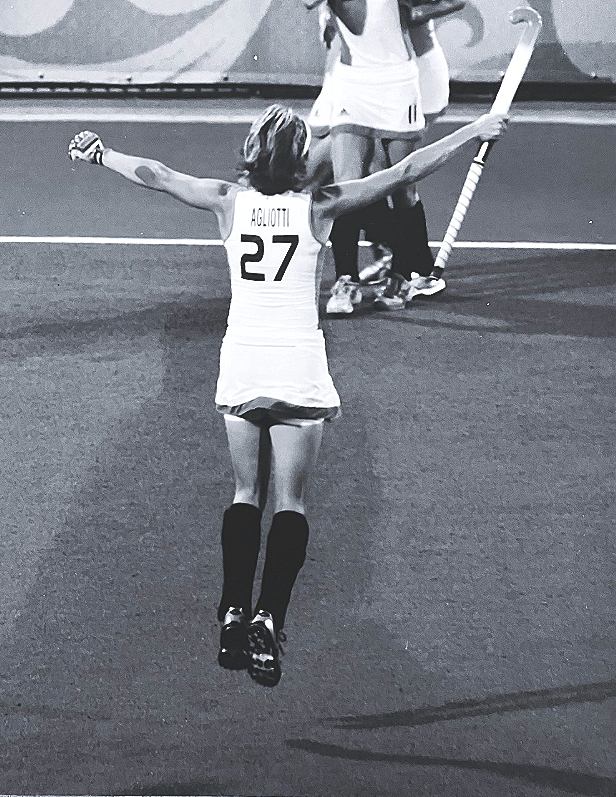 A female field hockey player wearing a jersey with the number 27 is celebrating with arms outstretched on a field. She is holding a hockey stick in her right hand and has black knee-high socks, with other players visible in the background.
