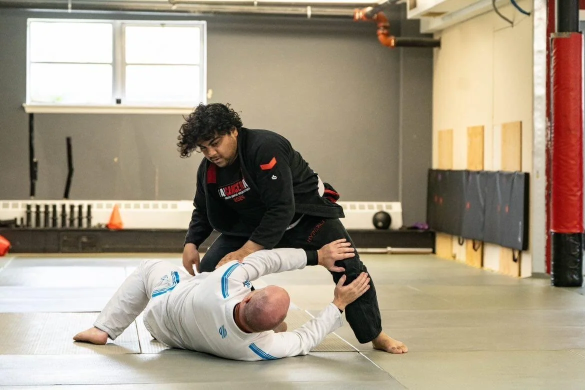 Two Brazilian Jiu-Jitsu practitioners training in a gym: one person is on the ground, and the other is standing with one knee on the ground, practicing a technique.