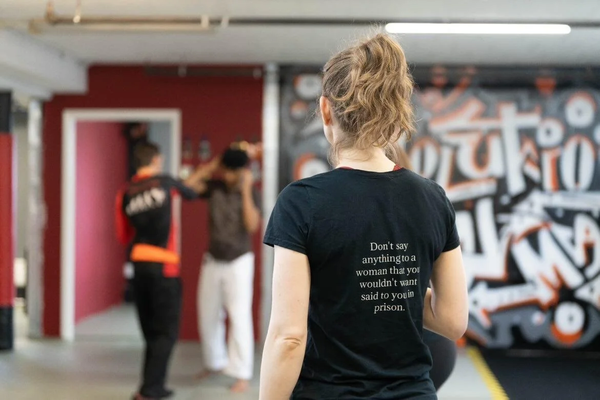A woman with curly hair wearing a black t-shirt with text on the back in a gym or boxing gym, with two people boxing in the background.