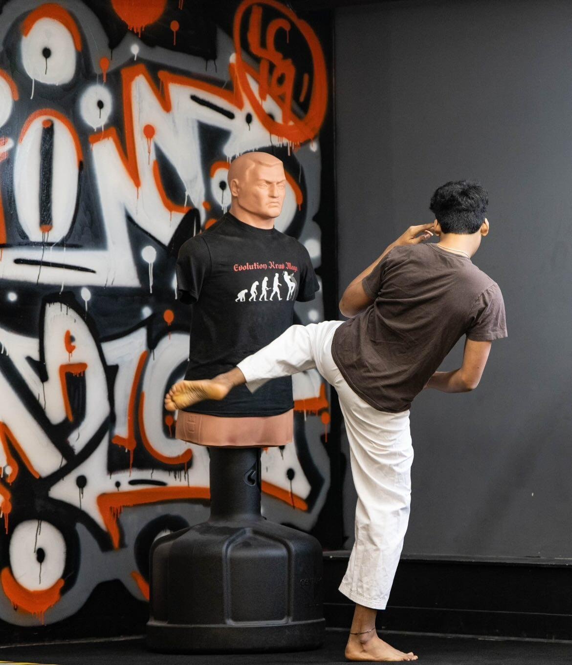A person practicing martial arts in front of a graffiti wall with a mannequin on a punching bag, the mannequin wears a black T-shirt with a graphic showing evolution and the words 'Evolution-Head Man.' The person is balancing on one leg with the other leg extended and resting on the mannequin's waist.