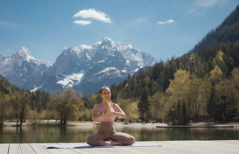A woman practicing yoga on a wooden platform near a lake with snow-capped mountains in the background. She is sitting cross-legged, smiling, with her hands in a prayer position.