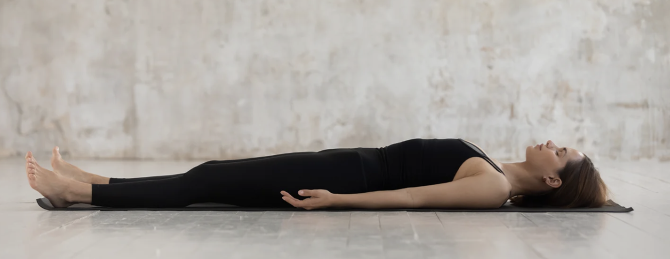 A woman in black workout clothes doing a relaxation pose on a yoga mat in a spacious room with a light-colored brick wall.