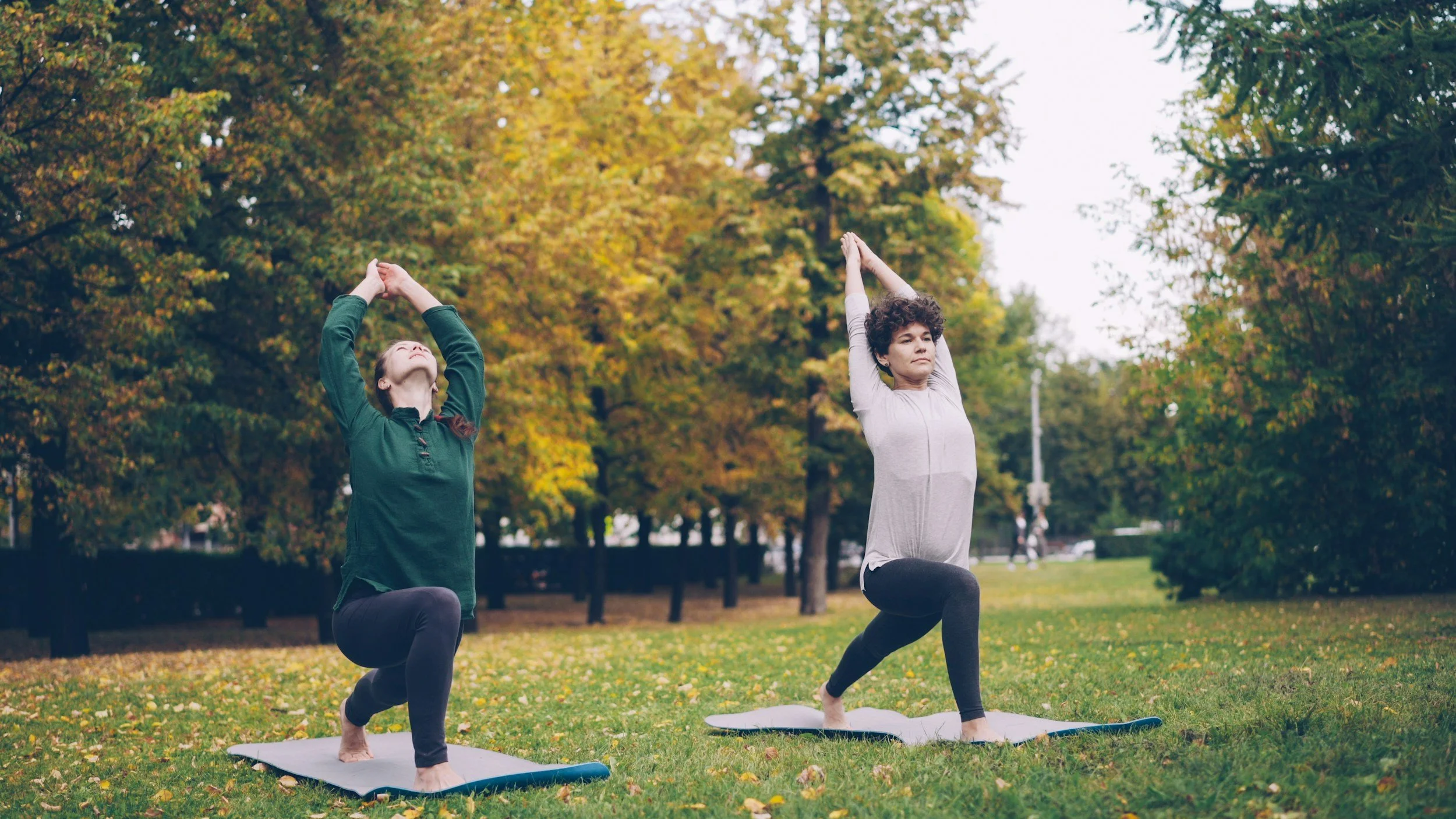 Two women practicing yoga outdoors in a park during fall, kneeling on yoga mats, stretching upwards with hands together, surrounded by trees with yellow and green leaves.