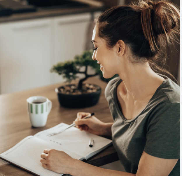 A woman with brown hair tied in a bun, smiling and writing in a notebook at a wooden table with a cup and a potted bonsai plant