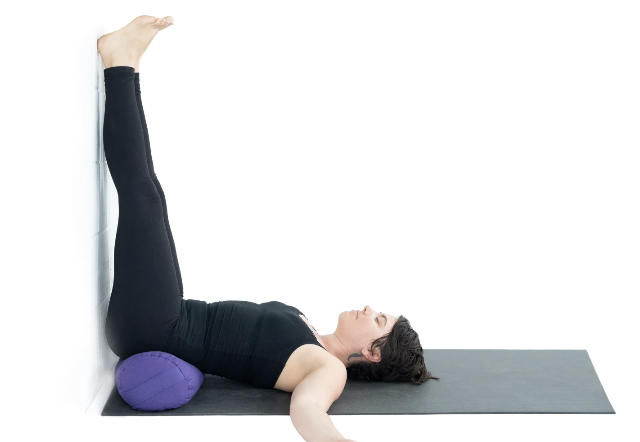 Woman practicing yoga or stretching on a mat with a purple bolster under her lower back, her legs raised against the wall.