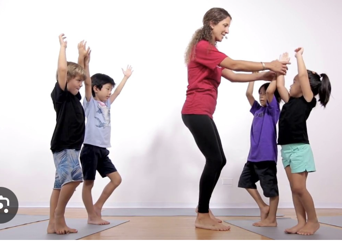 A woman leading a group of children in a fun activity, holding hands and raising them high, indoors with a white wall background.