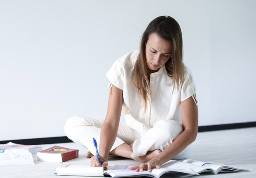 Woman sitting on floor, reading and writing in notebooks with books and papers scattered around.