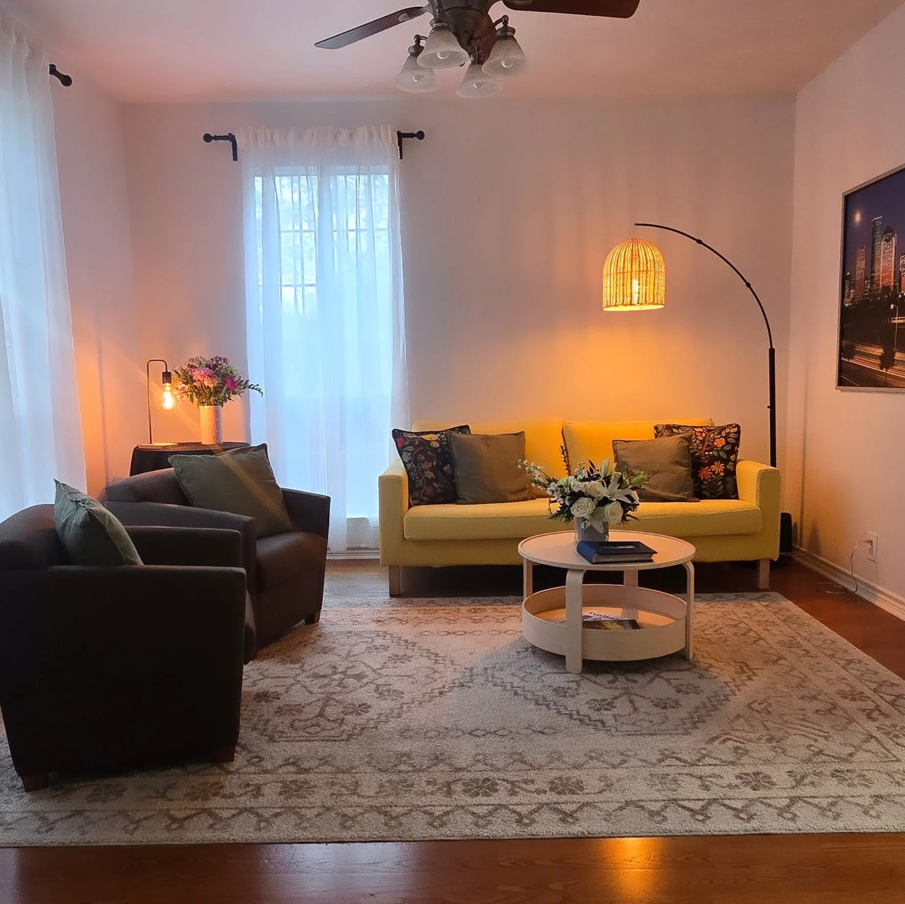 A living room with a dark sofa with pillows, a yellow sofa with decorative pillows, a white oval coffee table with magazines on it, and a rug with a floral pattern. There is a floor lamp with a curved arm and a basket-shaped lampshade, a table with a