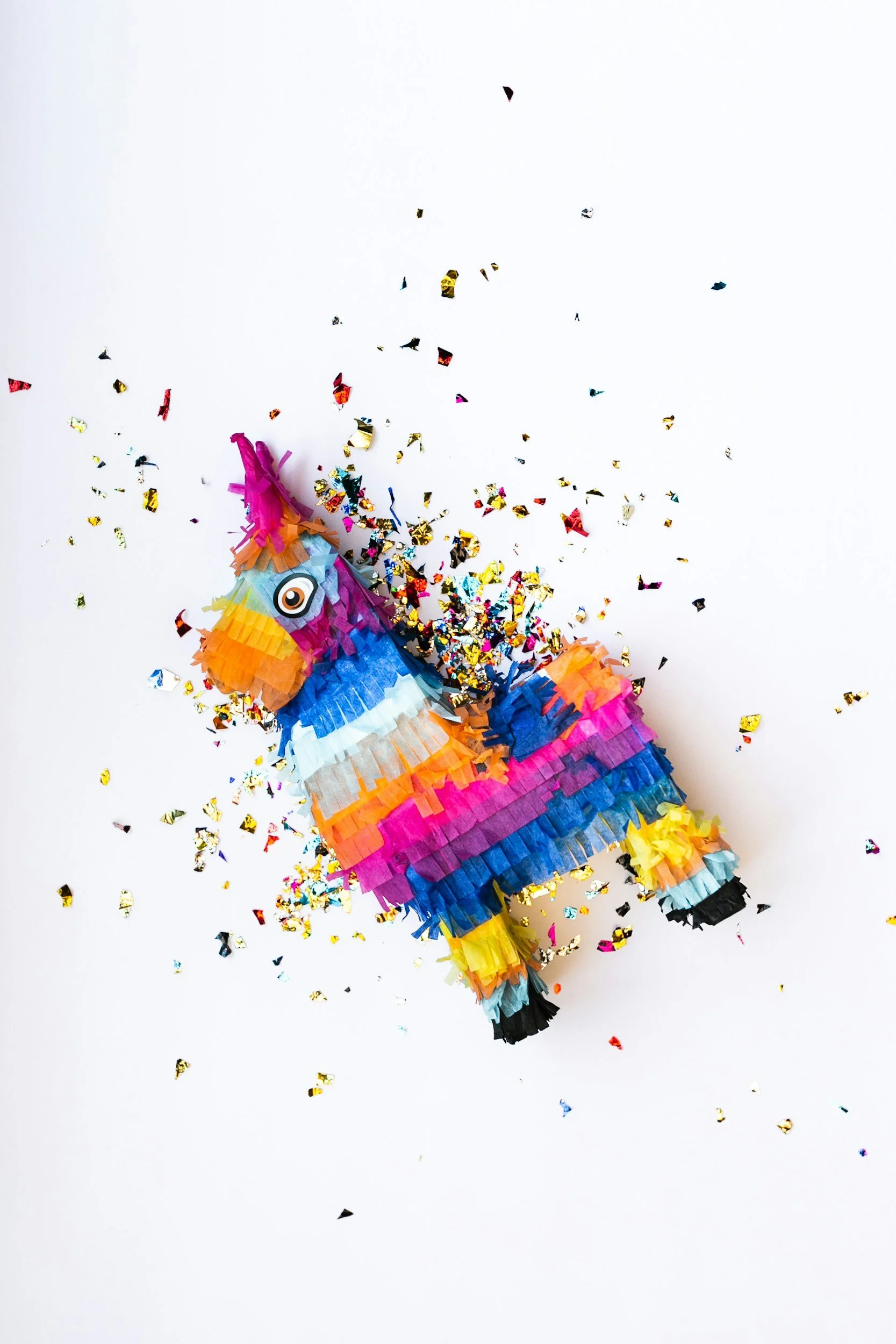 Colorful birthday piñata shaped like a unicorn with rainbow colors, surrounded by metallic confetti on a white background.