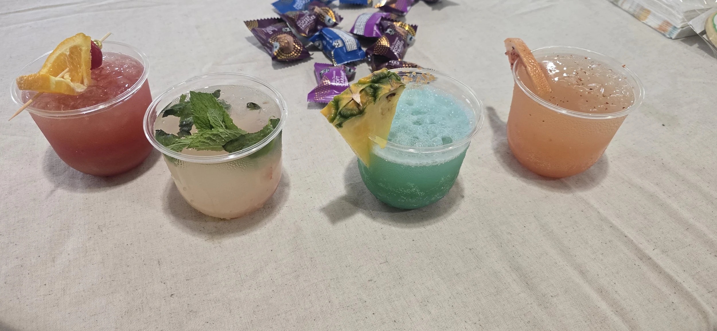 Four colorful drinks with garnishes on a table with candy wrappers in the background.