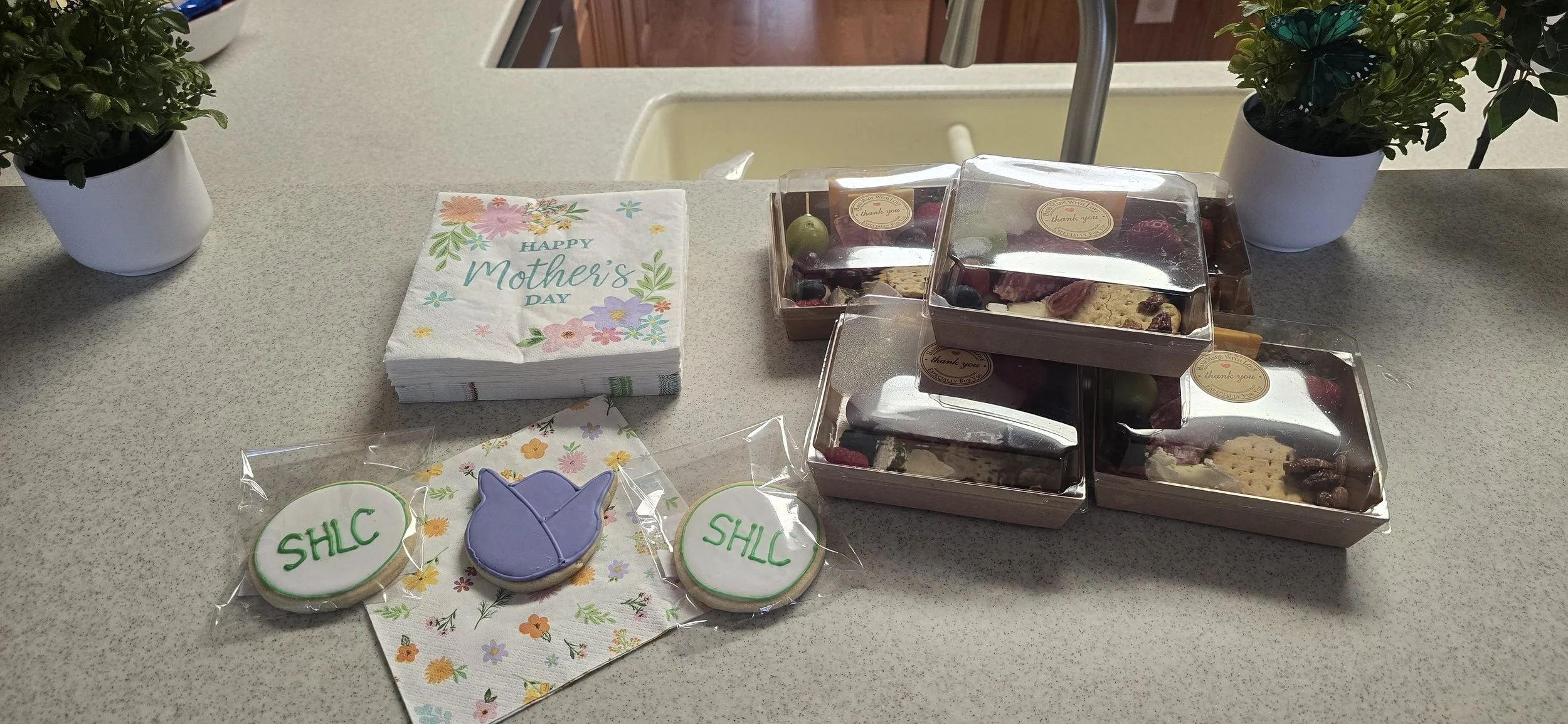 A countertop decorated for Mother's Day with a stack of paper napkins reading "Happy Mother's Day," a small gift bag with cookies or chocolates inside, some decorated cookies with