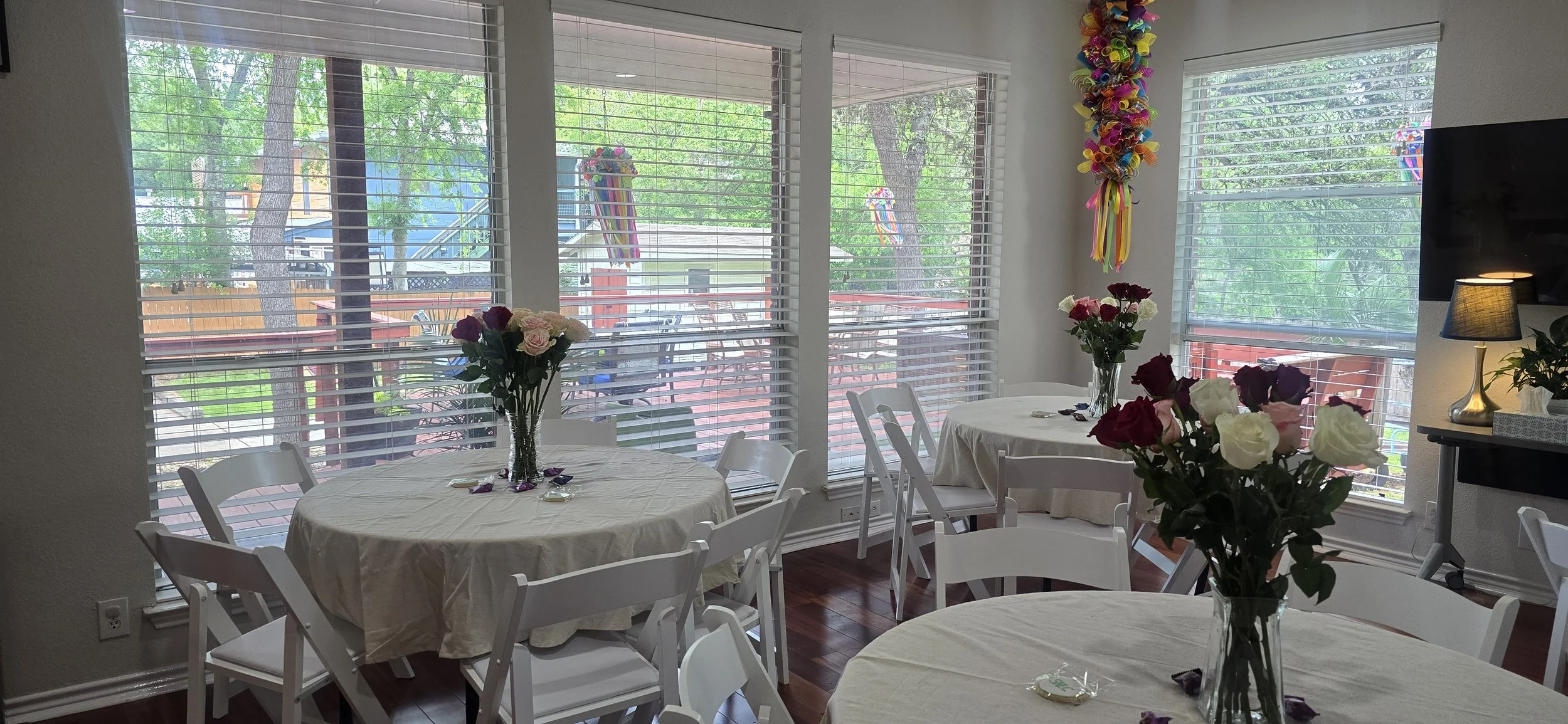 A decorated dining room with round tables covered in white tablecloths, each with a vase of pink and white roses. Large windows with blinds let in natural light. Colorful decorations hang from the ceiling, and a standing lamp is visible in the corner