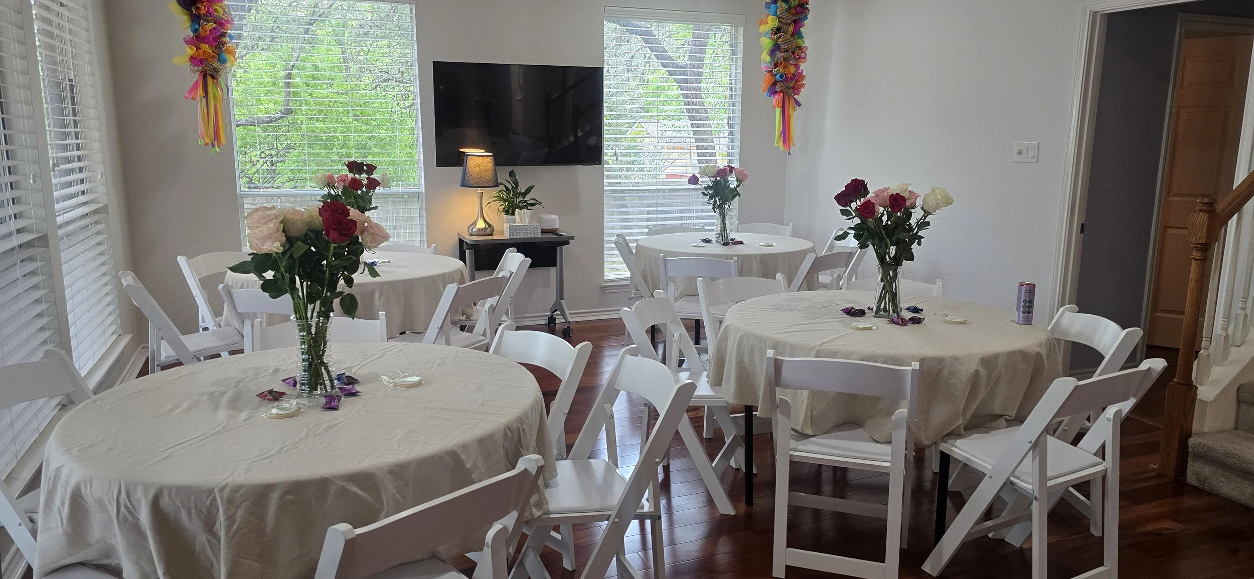 A dining room set up for a celebration with four round tables covered in beige tablecloths and decorated with glass vases of pink and white roses, surrounded by white folding chairs. There is a television mounted on the wall and a small black table w