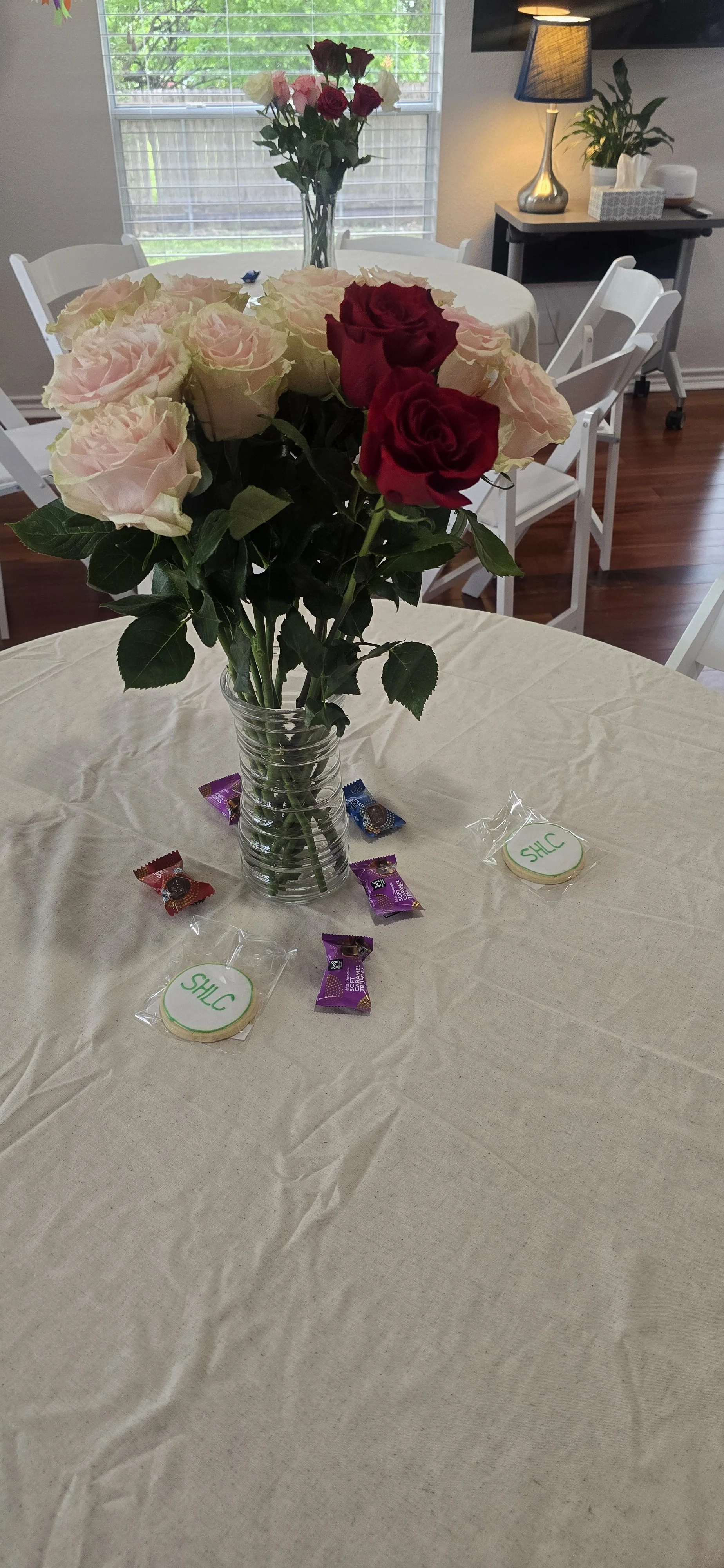 A dining table with a clear glass vase holding pink and red roses, and small chocolates and cookies with a SHLC label around the table. In the background, a window with blinds, and a side table with a lamp and plants.