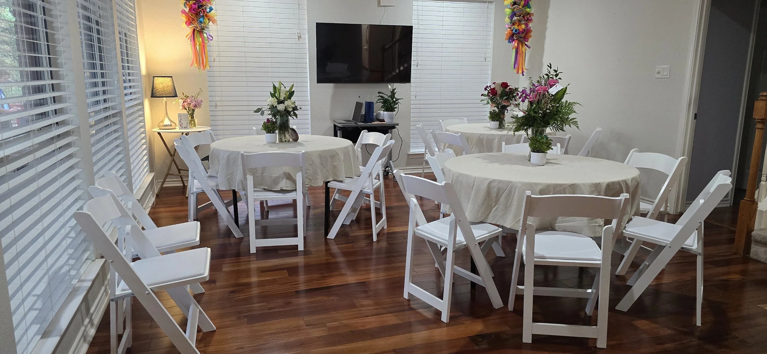 A dining area with three round tables covered with white tablecloths, decorated with floral centerpieces, surrounded by white folding chairs. The room features wooden flooring, white blinds covering the windows, a small side table with a lamp and vas