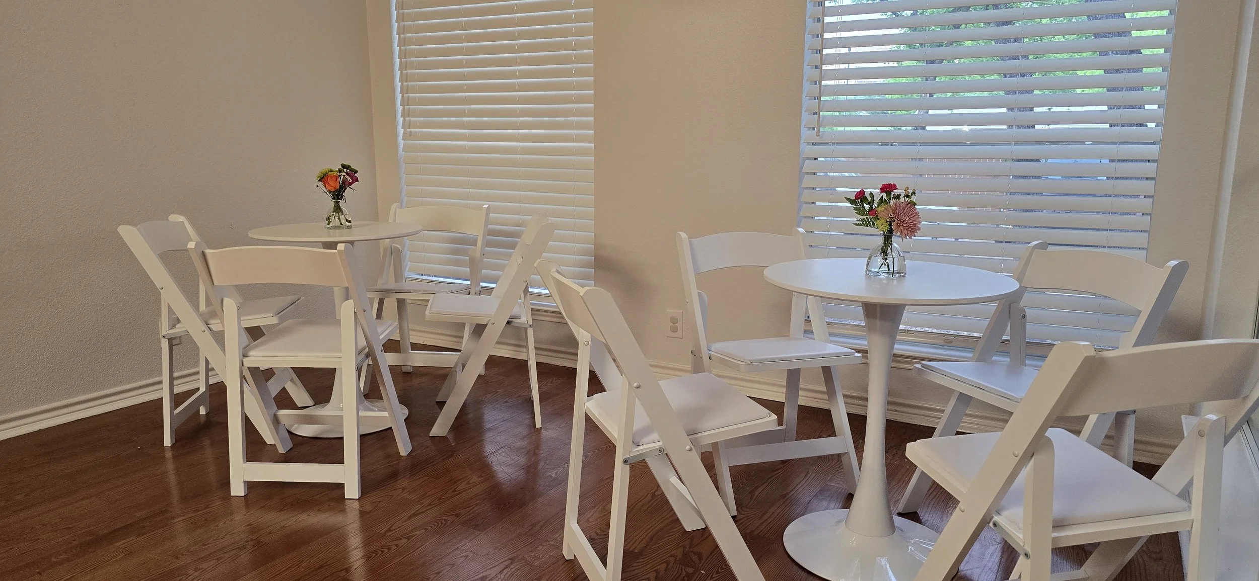 A cozy indoor space with white round tables and chairs, decorated with vases of colorful flowers, and large windows with white blinds letting in natural light.