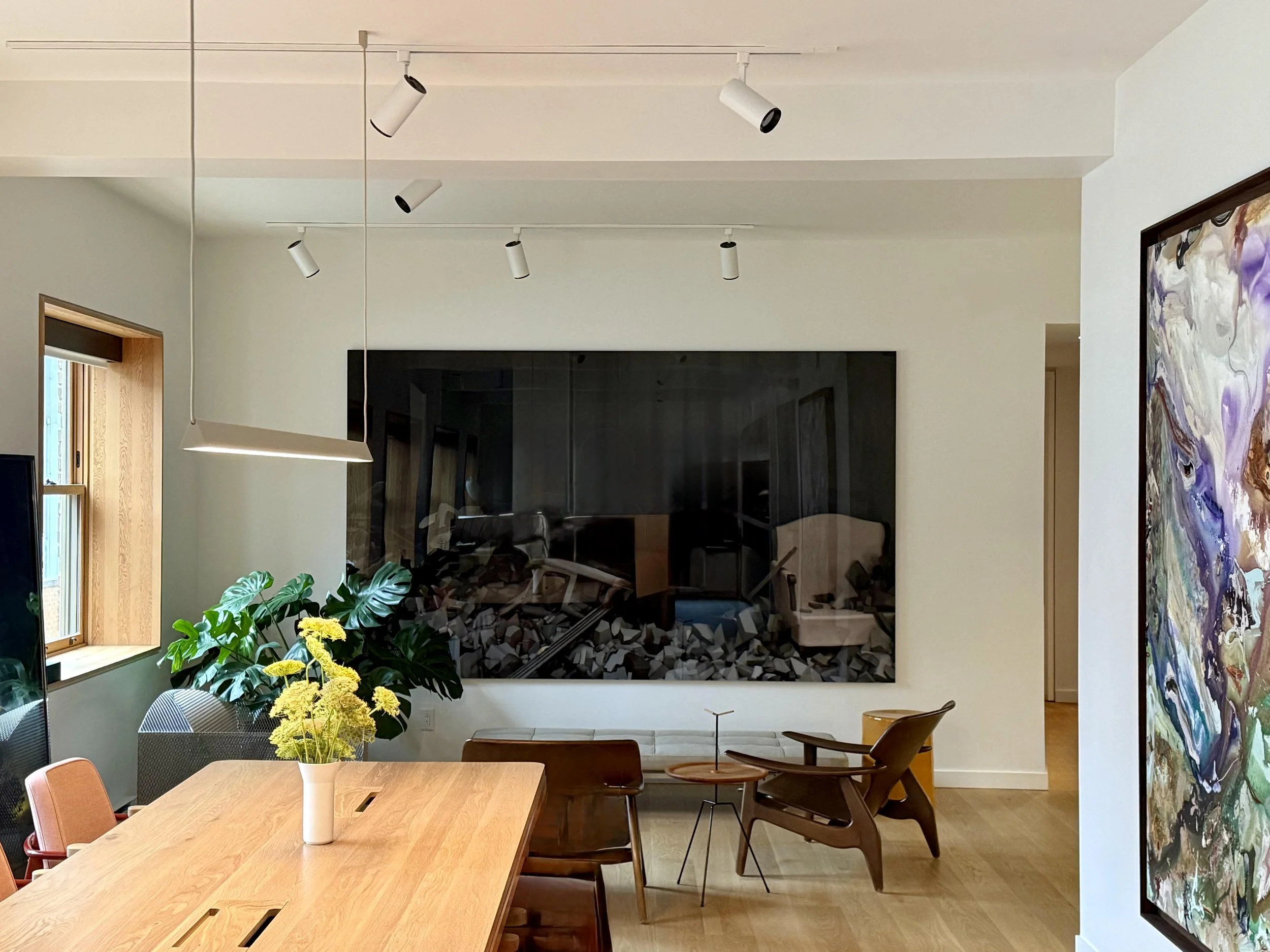 A modern dining room with a wooden table decorated with a white vase of yellow flowers, surrounded by orange, gray, and wooden chairs. A large window with a wooden frame lets in natural light. In the background, there is a framed abstract painting on the wall, a white ceiling with multiple adjustable track lights, and a contemporary black fireplace with stacked firewood underneath.