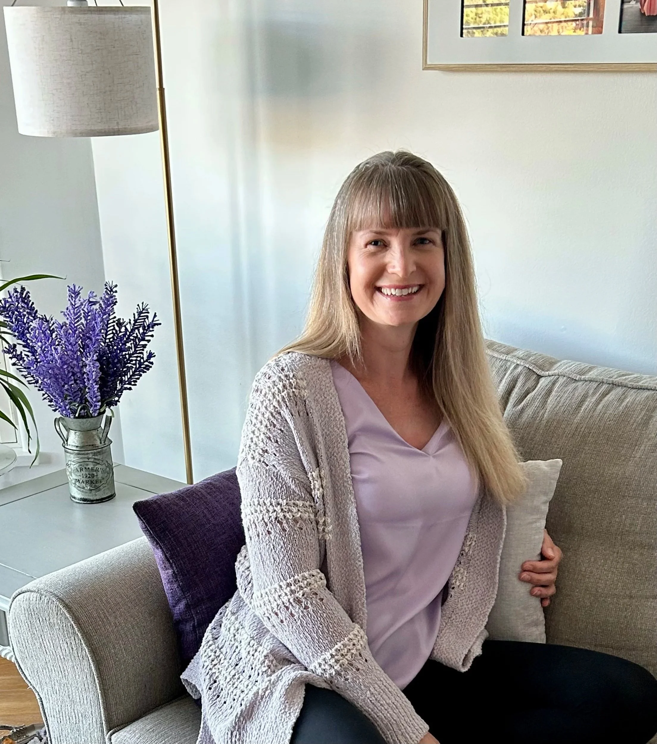 A woman with long blonde hair and a big smile sitting on a light-colored sofa in a living room. She is wearing a lavender top and a beige knitted cardigan. To her left, there is a white side table with a bouquet of purple flowers in a vintage-style watering can. Behind her, a beige cushion is visible. A tall floor lamp with a cream-colored shade and gold stand is positioned next to the sofa. The background features a white wall with a framed picture and part of a window showing autumn trees.