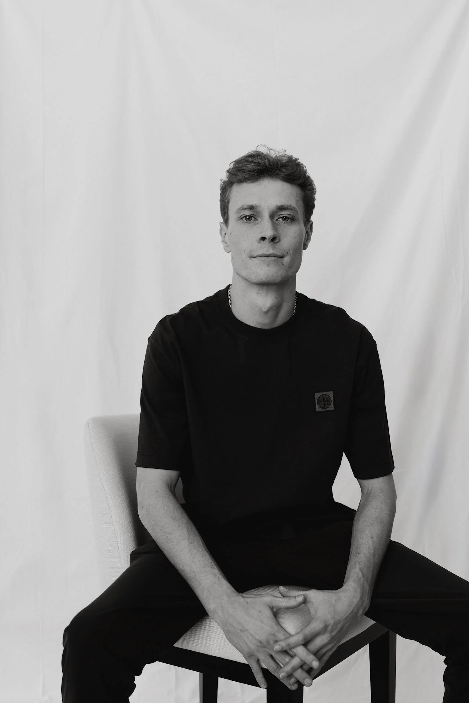 A black and white photo of a young man sitting on a chair, with his hands clasped on his lap, wearing a black t-shirt, against a plain backdrop.