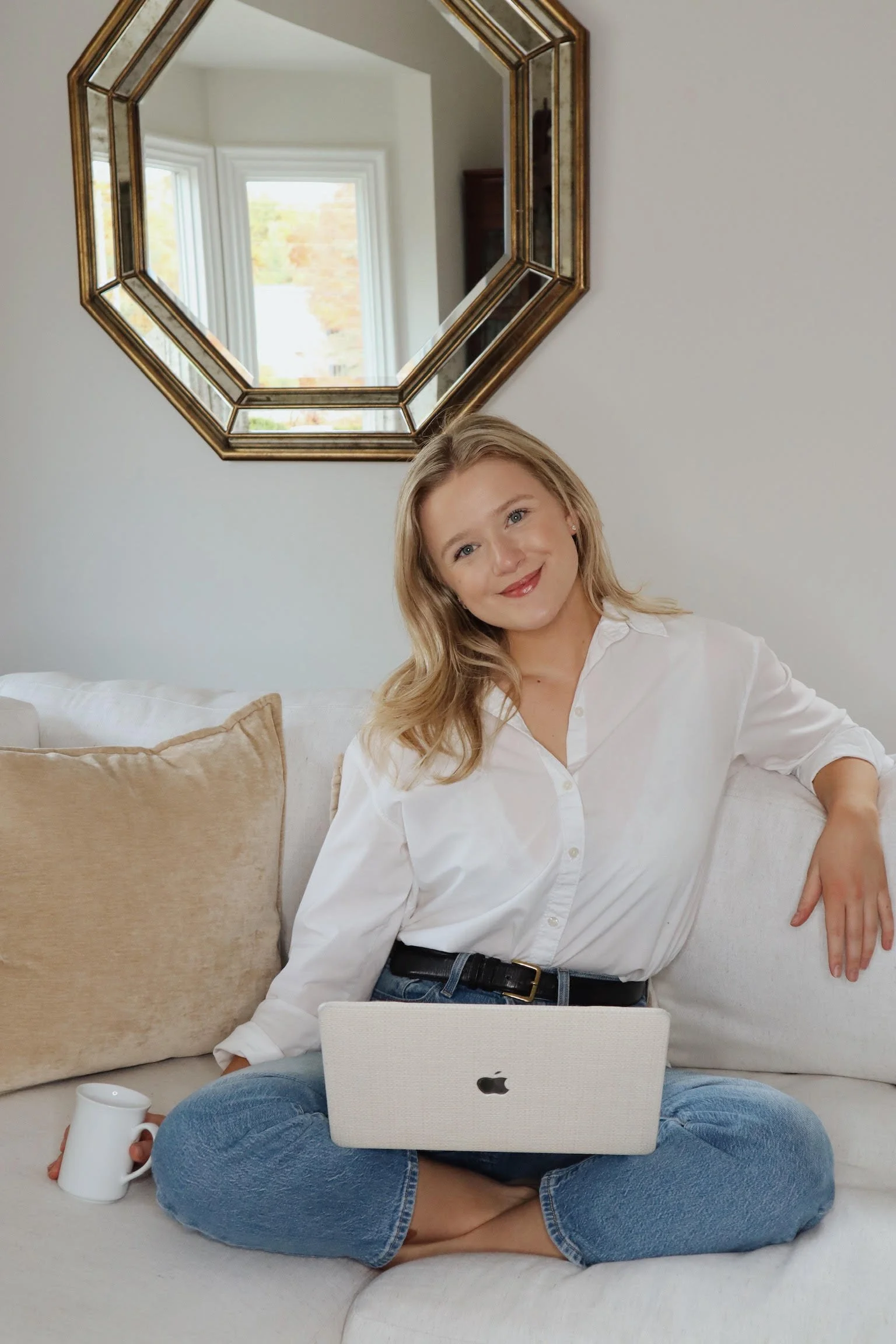 A woman with blonde hair smiling and sitting cross-legged on a light-colored sofa with beige cushions, using a silver Apple MacBook on her lap. She is holding a white mug in her left hand and is wearing a white button-up shirt and blue jeans. Behind her, there is a wall with a large octagonal mirror and a window reflecting trees and a brick wall.