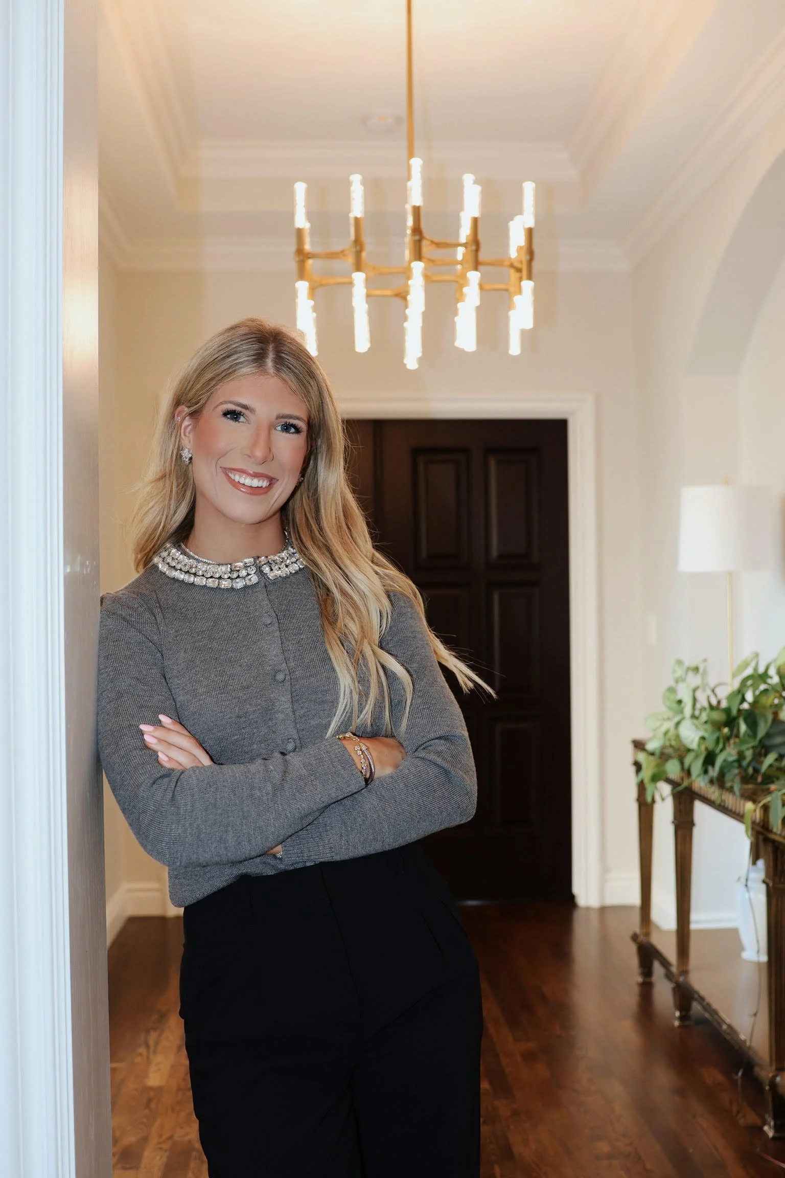 A woman with blonde hair and a smile, dressed in a gray top with a jewel neckline and black skirt, posing with arms crossed in a well-lit hallway with a chandelier and wooden flooring.