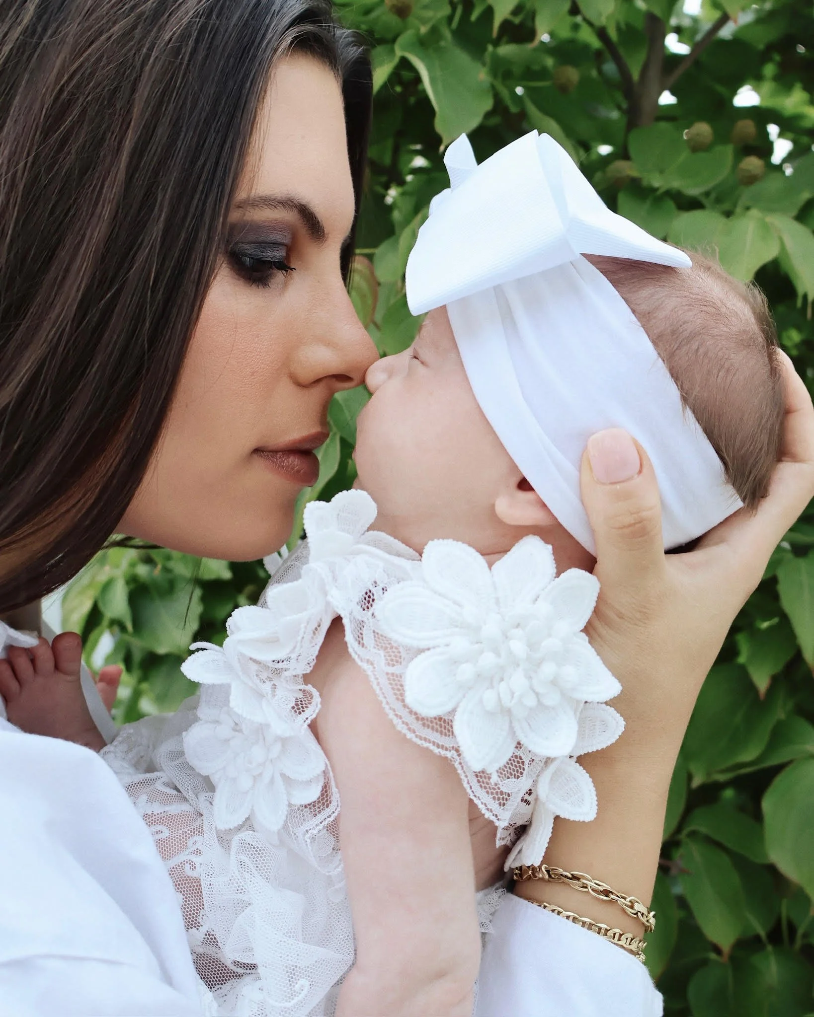 A woman with dark hair and makeup holds a baby with a white headband and dress, touching their noses together outdoors among green leaves.
