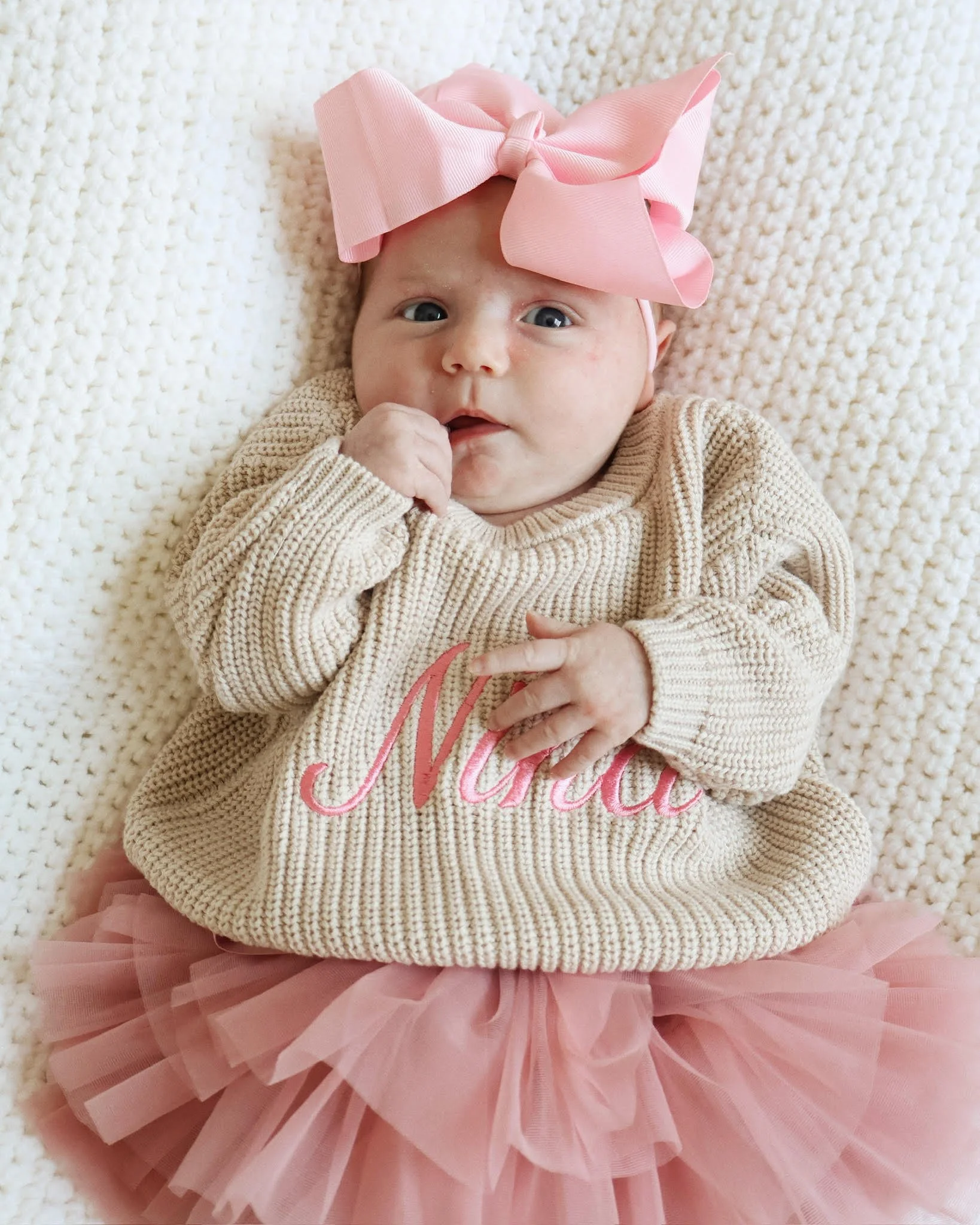 A baby girl lying on a soft, cream-colored blanket, wearing a beige knitted sweater with pink embroidery, pink tulle skirt, and a large pink bow headband.