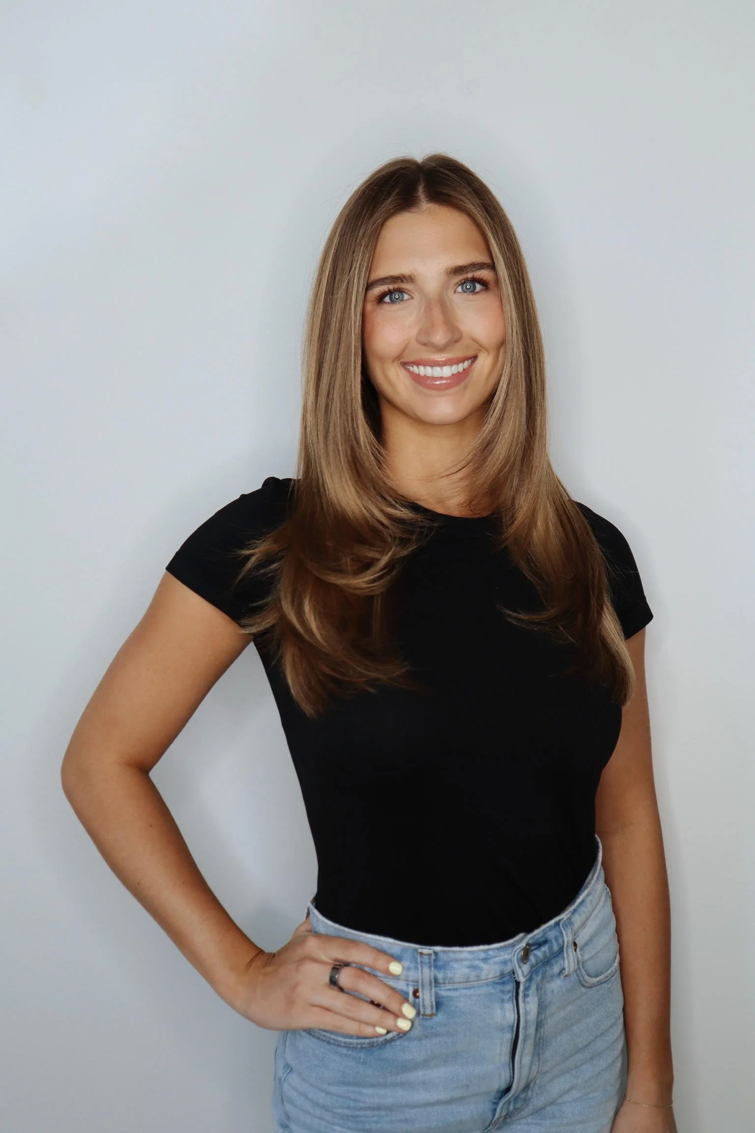 A young woman with long, wavy brown hair, blue eyes, and a bright smile standing against a plain light-colored wall. She is wearing a black t-shirt and light blue jeans, with one hand on her hip, posing confidently.