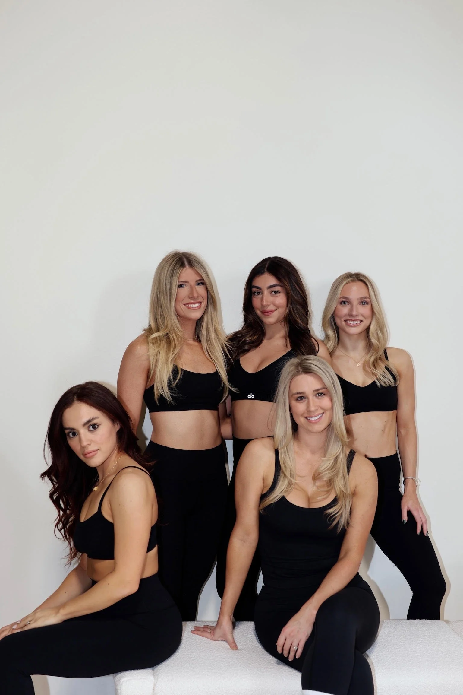 Group of six women in black workout clothing posing together indoors against a plain white wall.