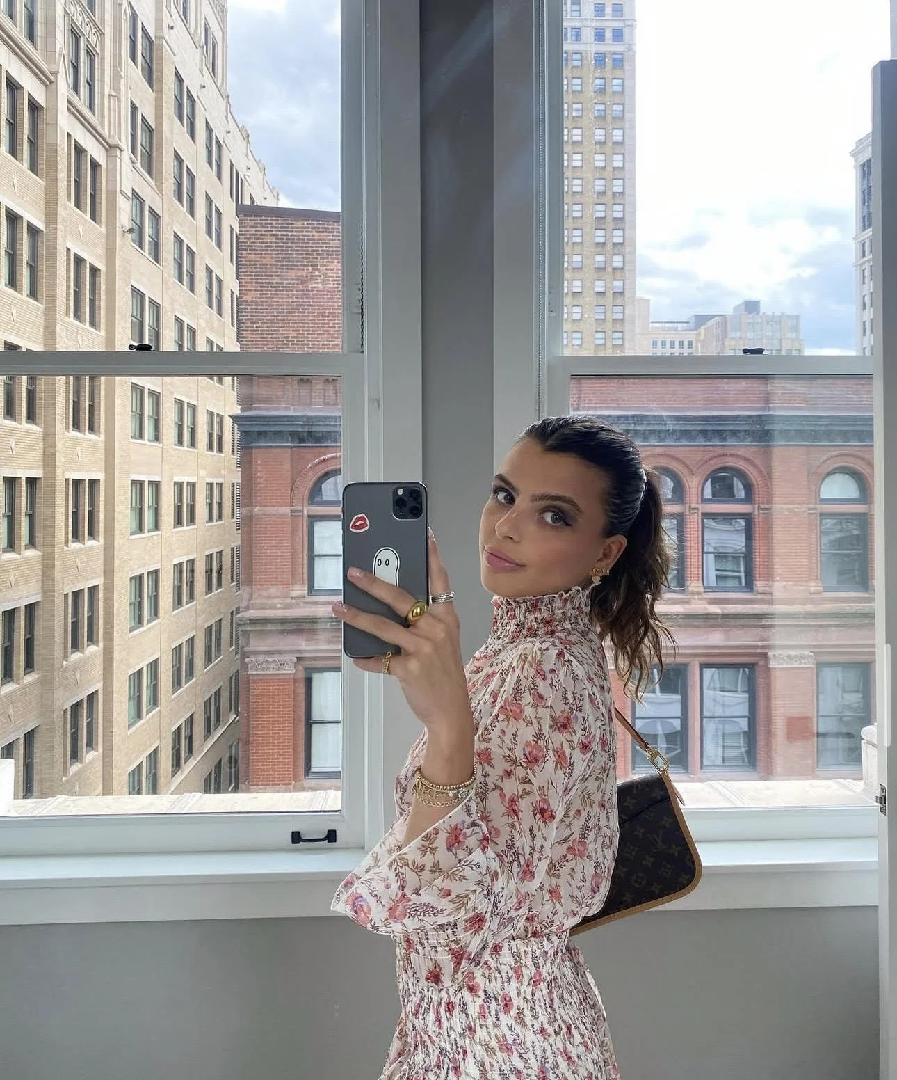 A woman takes a mirror selfie near a large window showing city buildings. She has dark hair tied back, wears a floral dress, and carries a designer handbag.
