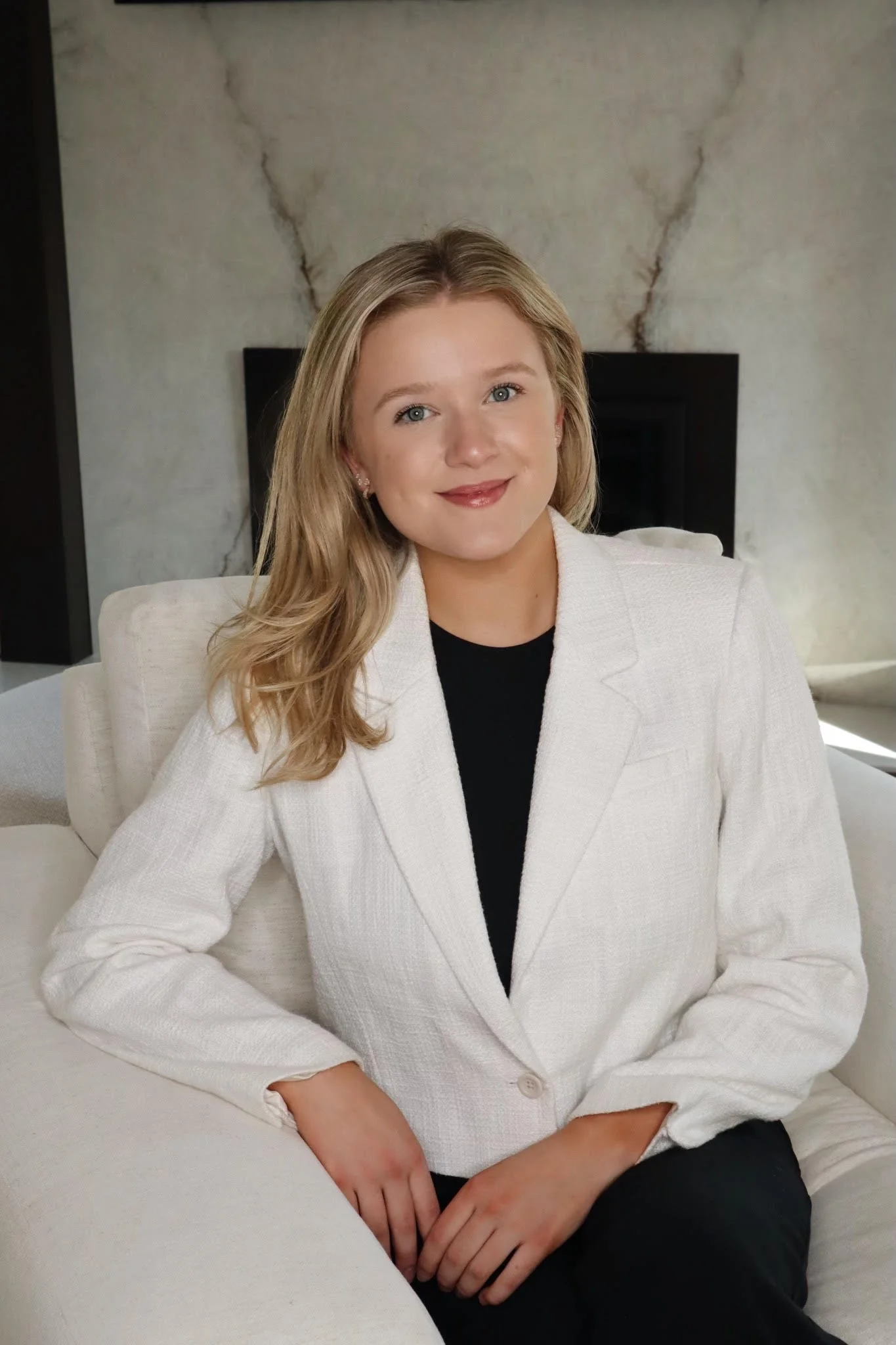 A woman with blonde hair wearing a white blazer sits on a white chair with her hands resting on her lap, in front of a marble wall.