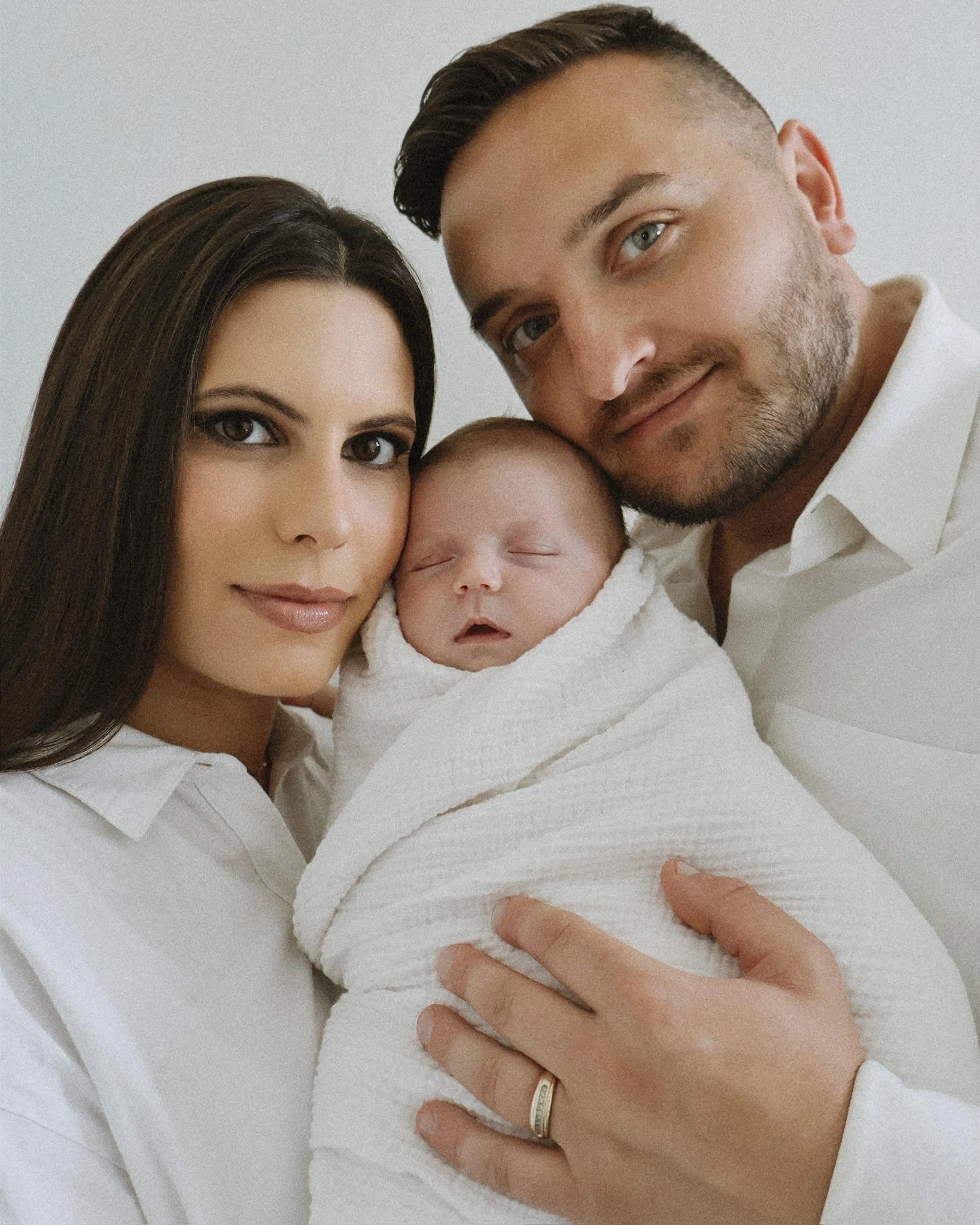 A woman with long dark hair and a man with short dark hair and a beard hold a sleeping baby wrapped in a white blanket. The woman wears a white top, and the man has a wedding ring and a white shirt. All are close together in an intimate moment, looking relaxed and peaceful.
