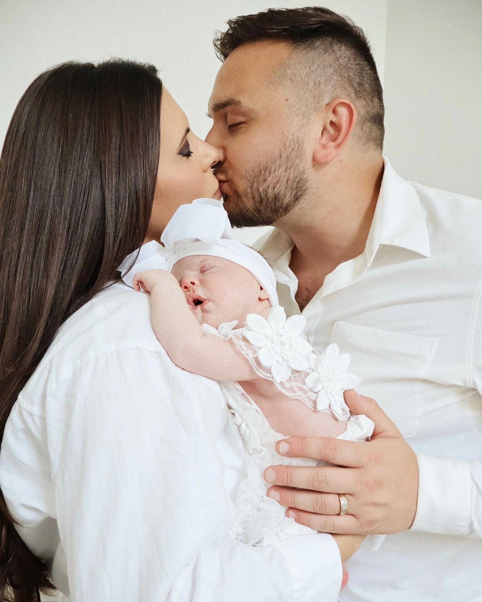 A couple sharing a kiss, holding a sleeping baby, all dressed in white, with a plain light background.