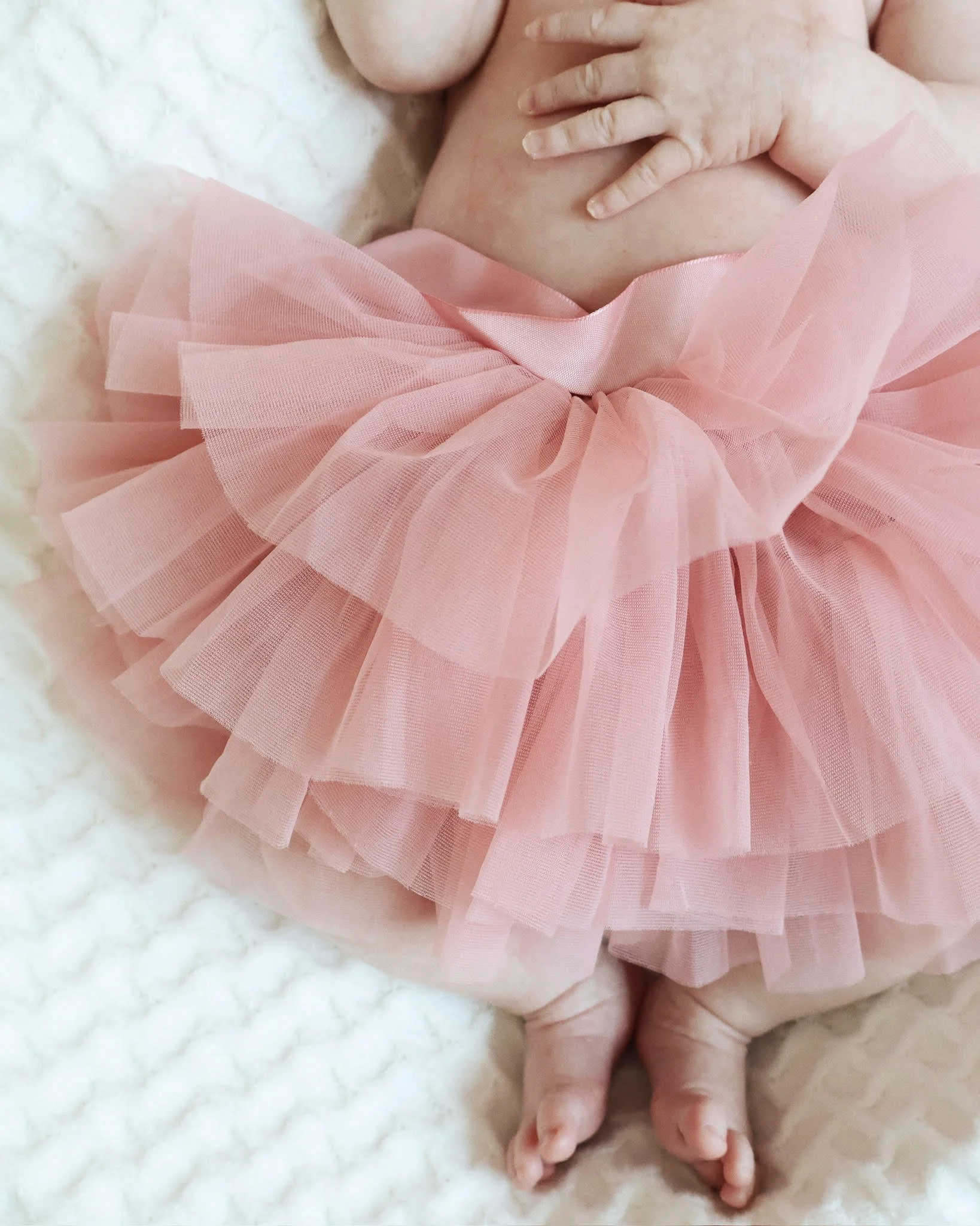 Close-up of a newborn baby's hand resting on a pink tulle skirt, lying on a soft white blanket.