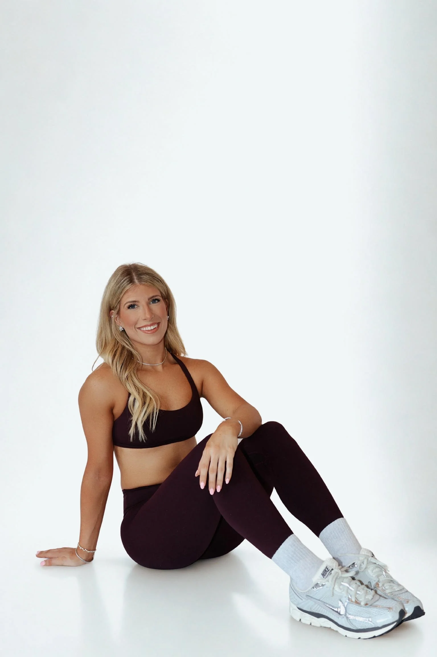 Smiling woman with blonde hair wearing a black sports bra, black leggings, gray Nike sneakers, and white socks, sitting on a clean white background.