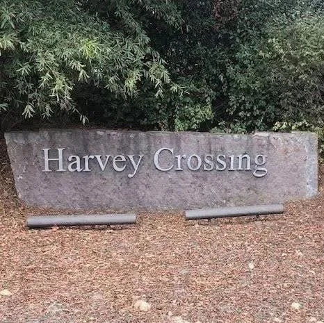A stone sign that reads "Harvey Crossing" with bushes and trees in the background.