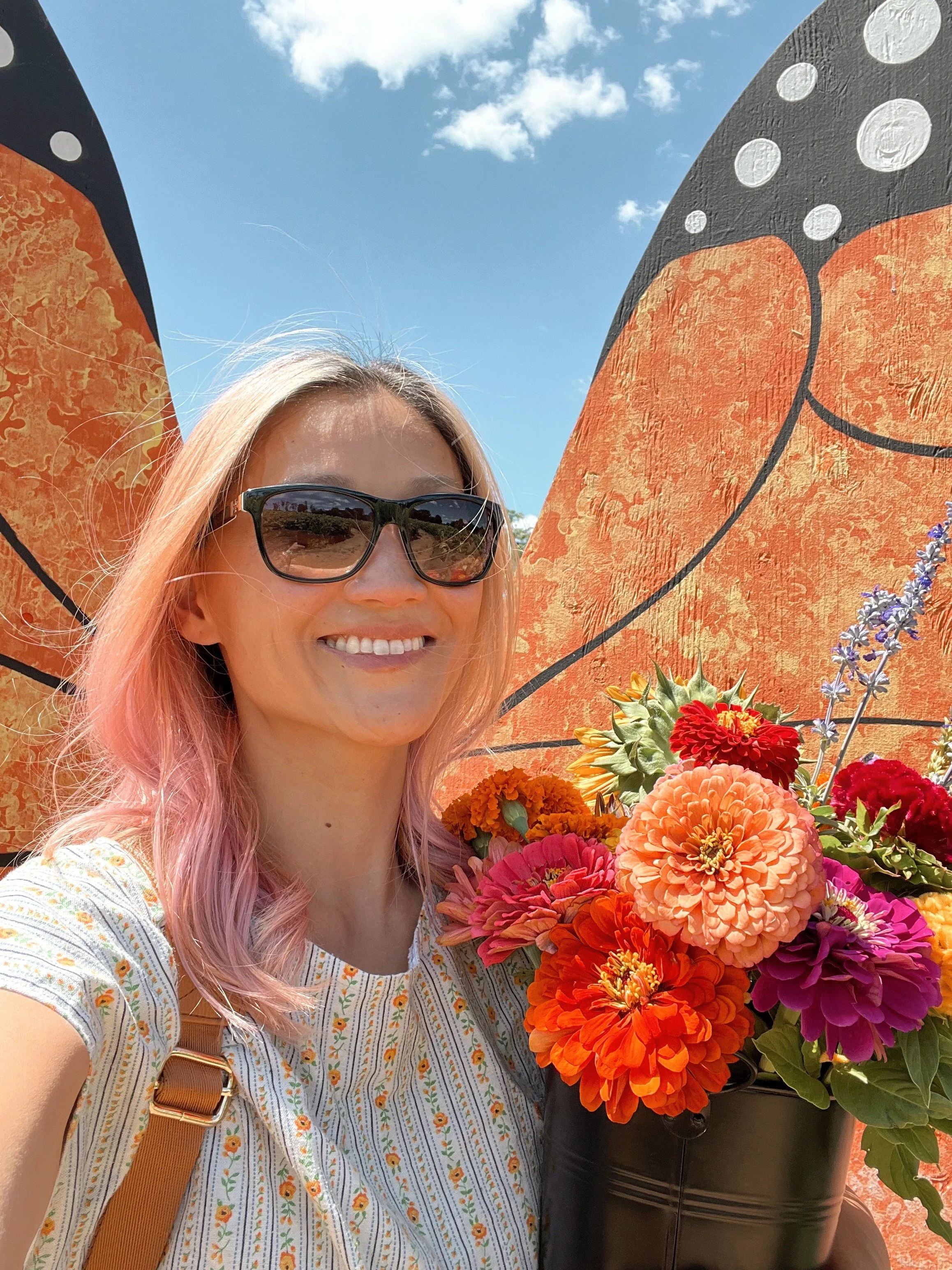 A young woman with pink hair wearing sunglasses and a white dress with yellow flowers, smiling while taking a selfie outdoors next to a large flower arrangement and a mural of butterfly wings painted on a wall.