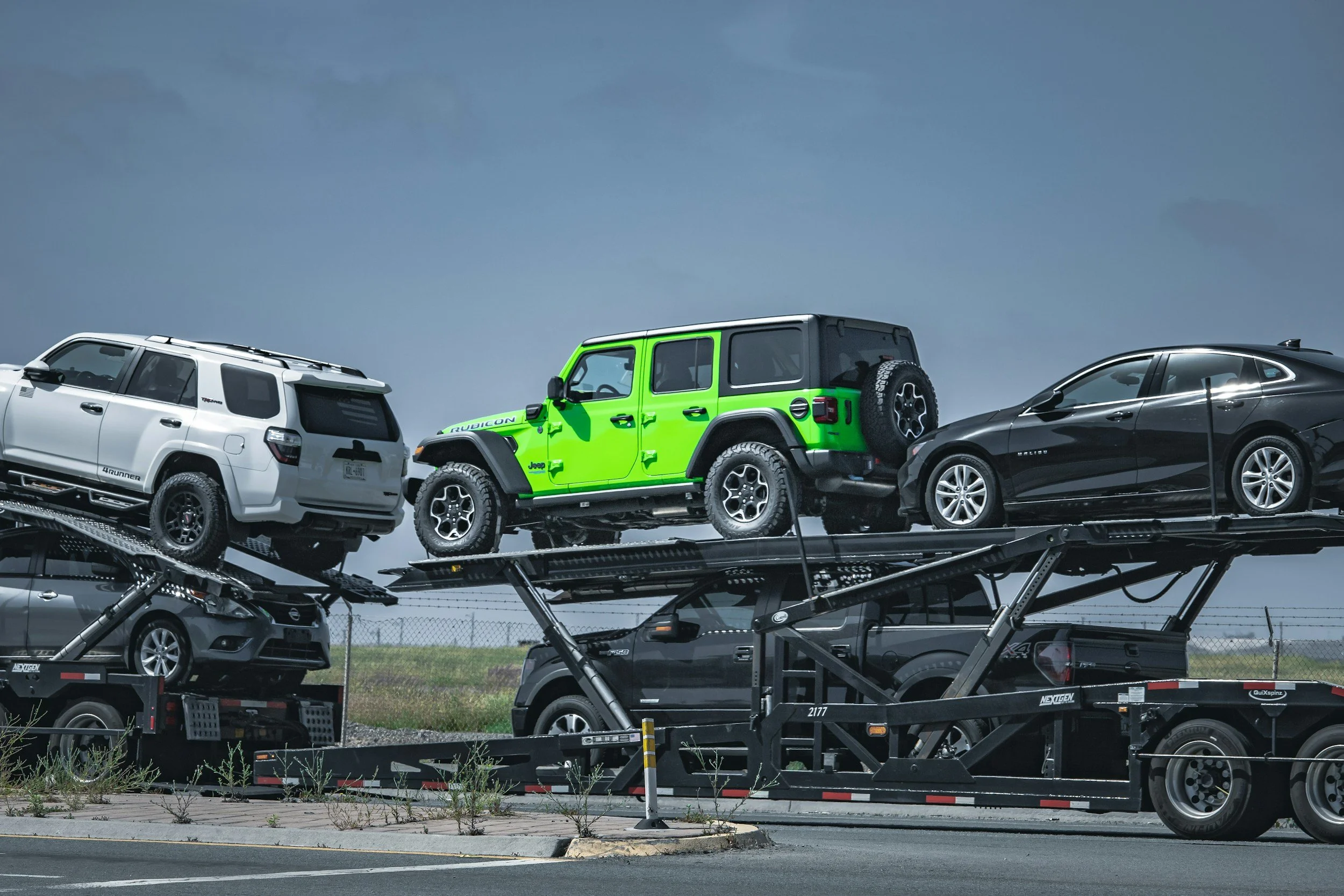 Multiple cars on a black car carrier trailer, including a white Jeep Rubicon and a black sedan, parked outdoors with a cloudy sky in the background.