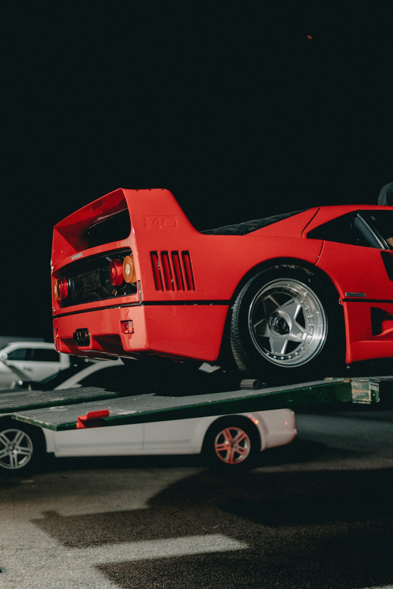 A red Ferrari sports car is being transported on a flatbed trailer at night, with a dark sky above and part of a car visible in the bottom foreground.